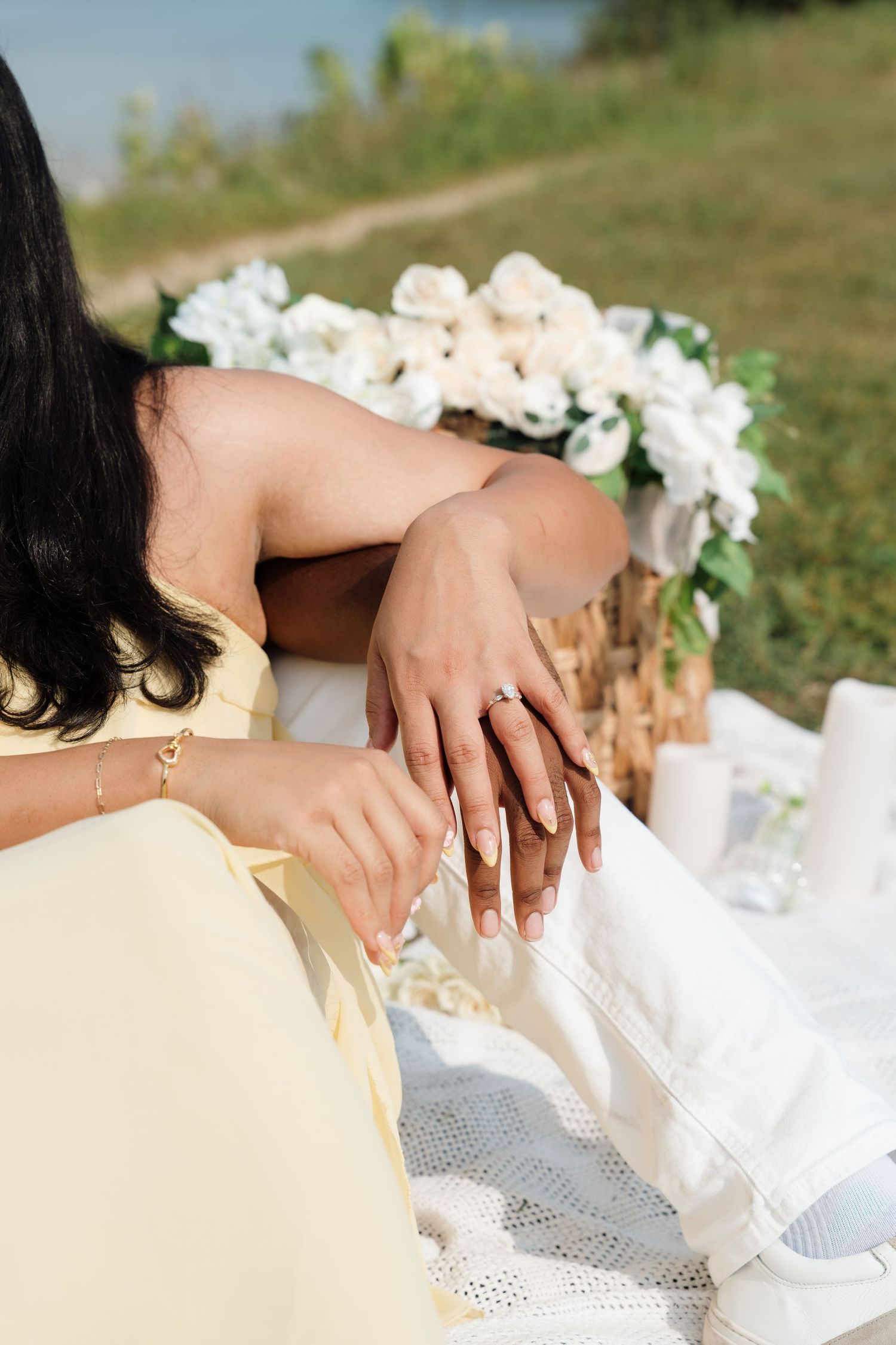 Woman in yellow dress showing engagement ring at outdoor picnic with white floral bouquet in background.