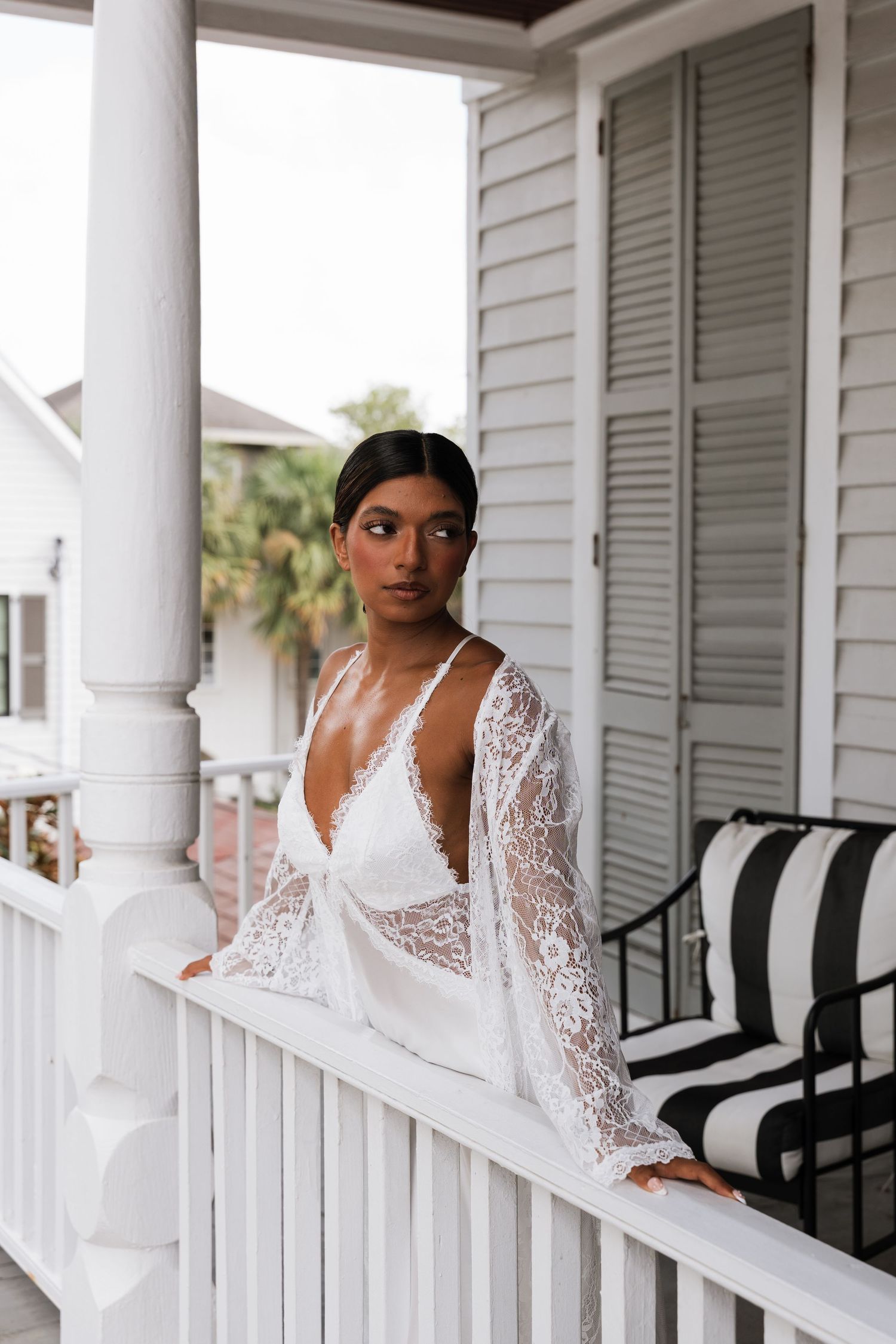 Woman in white lace bridal robe poses elegantly on a classic white porch with striped outdoor furniture.