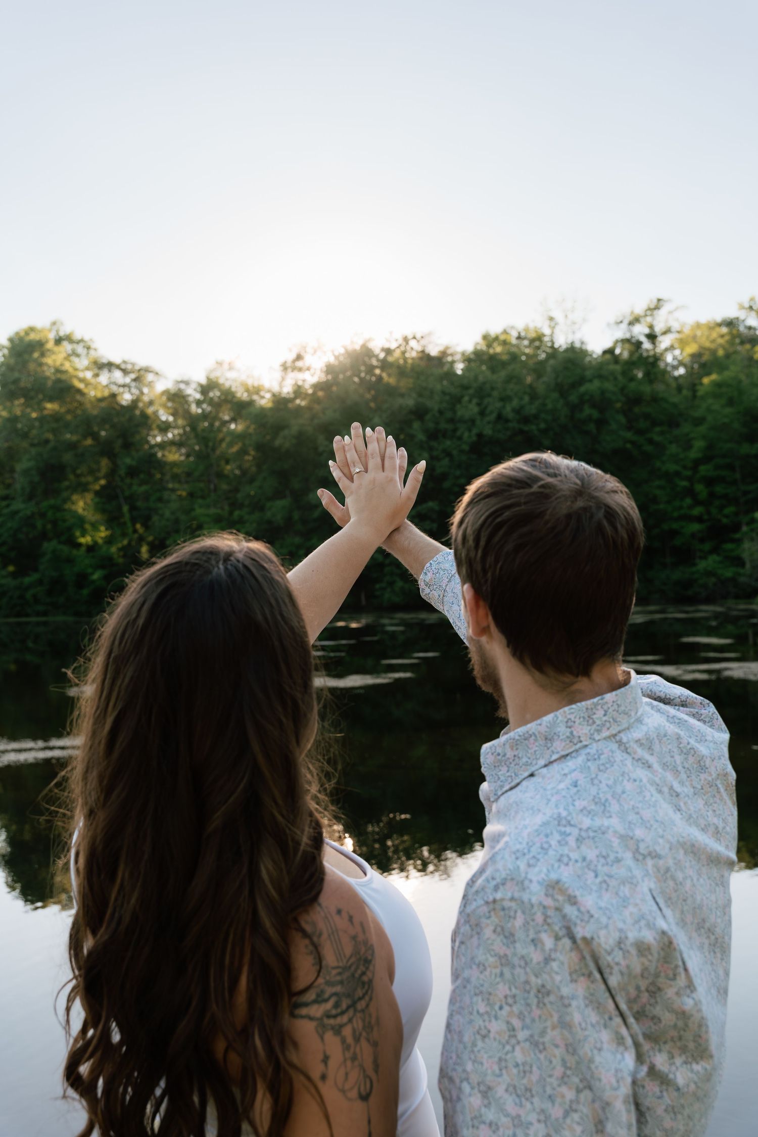 Couple holding hands up toward sunset over a reflective lake surrounded by lush green trees, woman with tattoo.