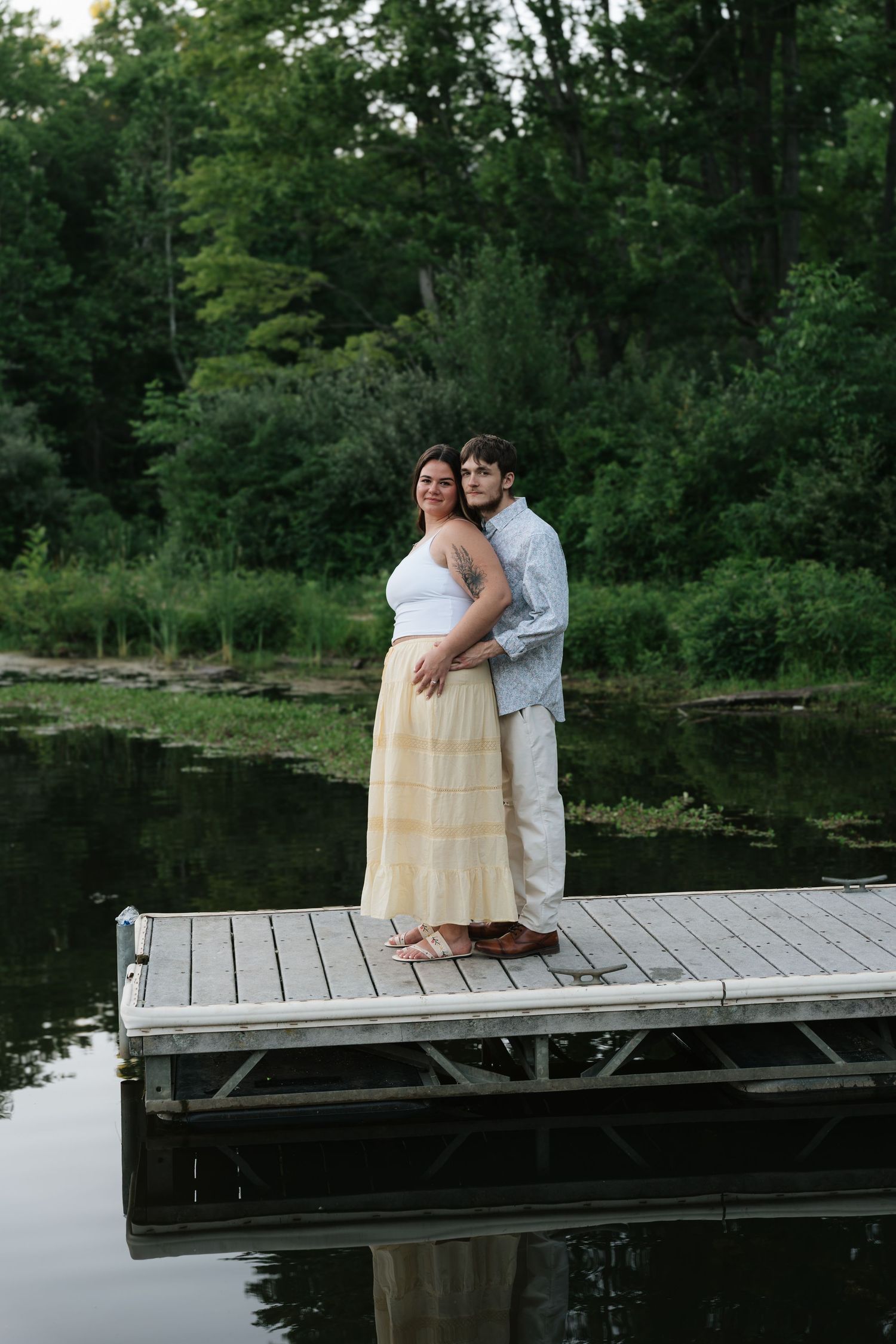 Couple embracing on a wooden dock by a serene pond surrounded by lush green trees.