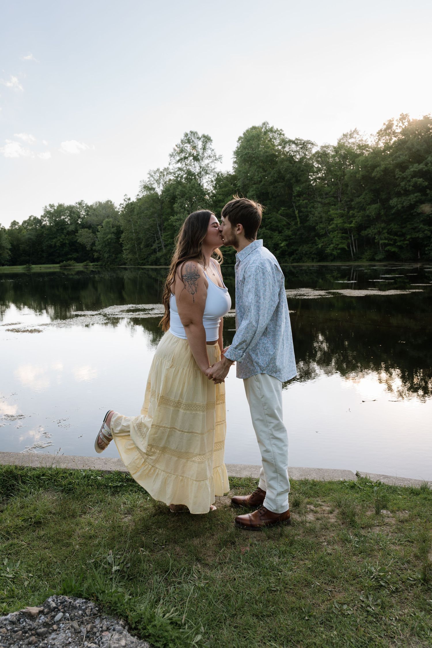 Couple kissing by a serene lake at sunset, woman in yellow skirt and white top, man in light blue shirt.