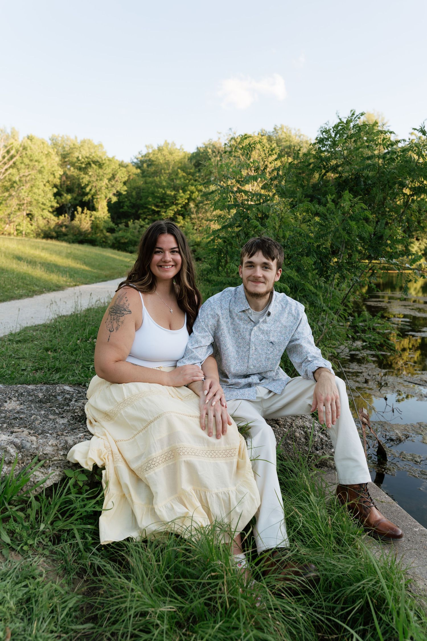 A smiling couple sits together on a rock near a pond, surrounded by lush green trees in a park setting.