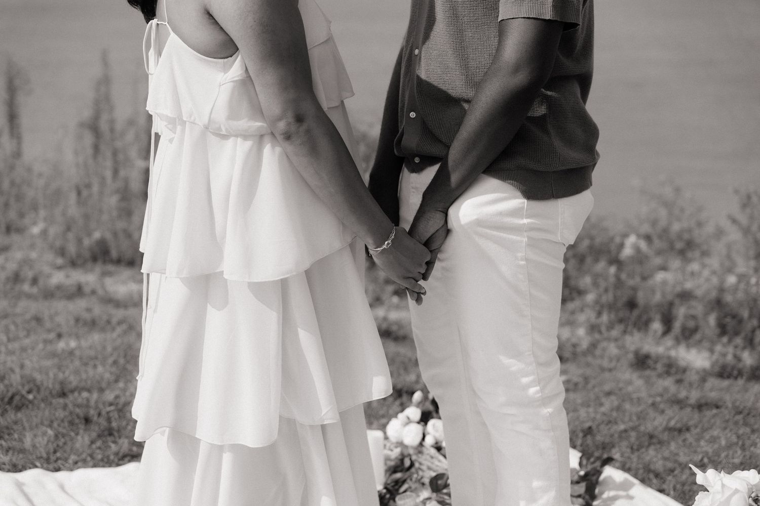Black and white photo of a couple holding hands, bride in tiered white dress and groom in light outfit outdoors.