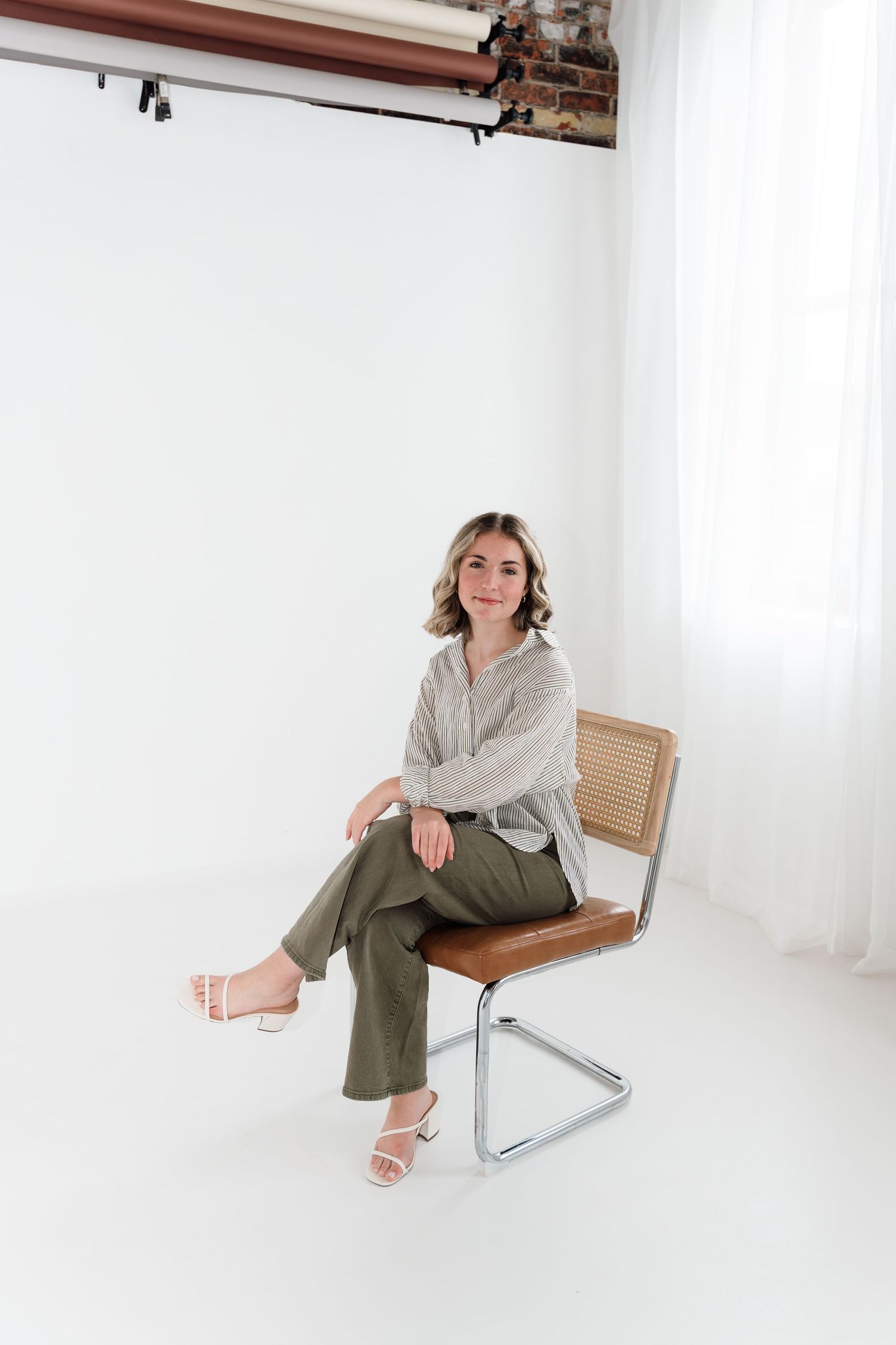 Woman in olive pants and striped blouse sitting on a modern cane chair in a white photography studio.