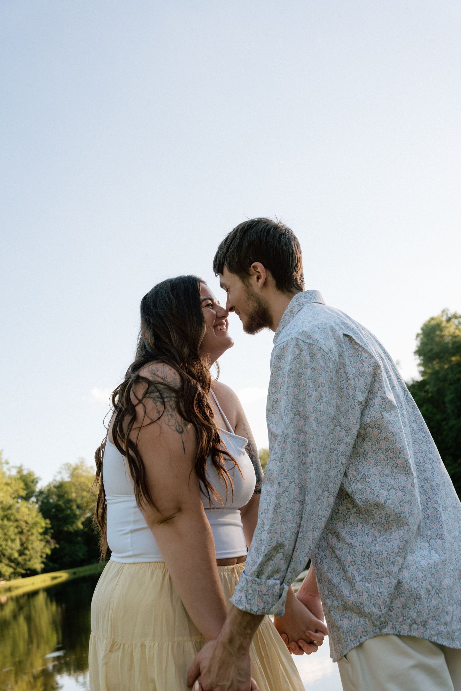Romantic couple holding hands and sharing a kiss outdoors near a lake on a sunny day.