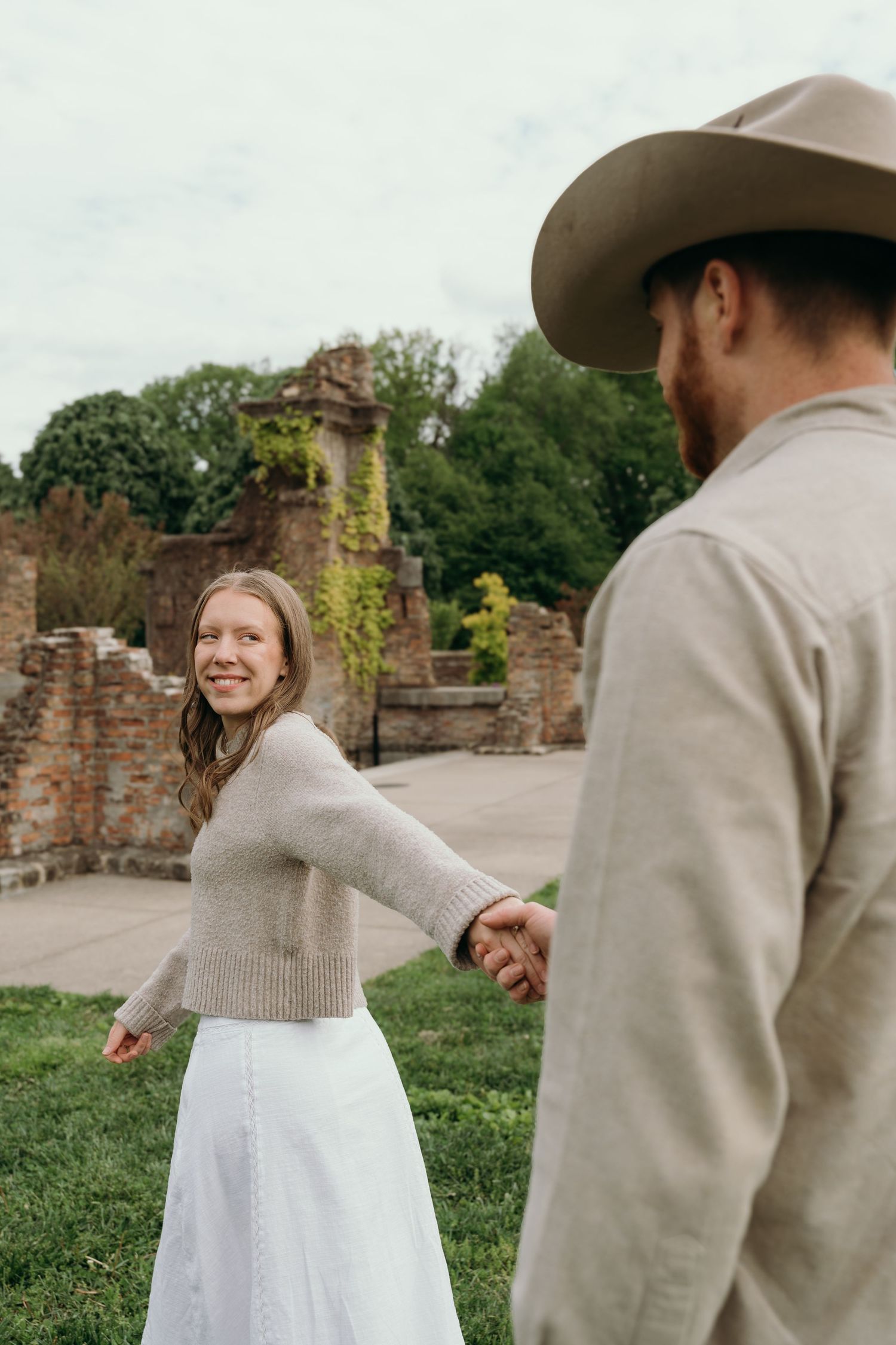 A smiling woman in a white skirt holds hands with a man in a cowboy hat near rustic brick ruins outdoors.