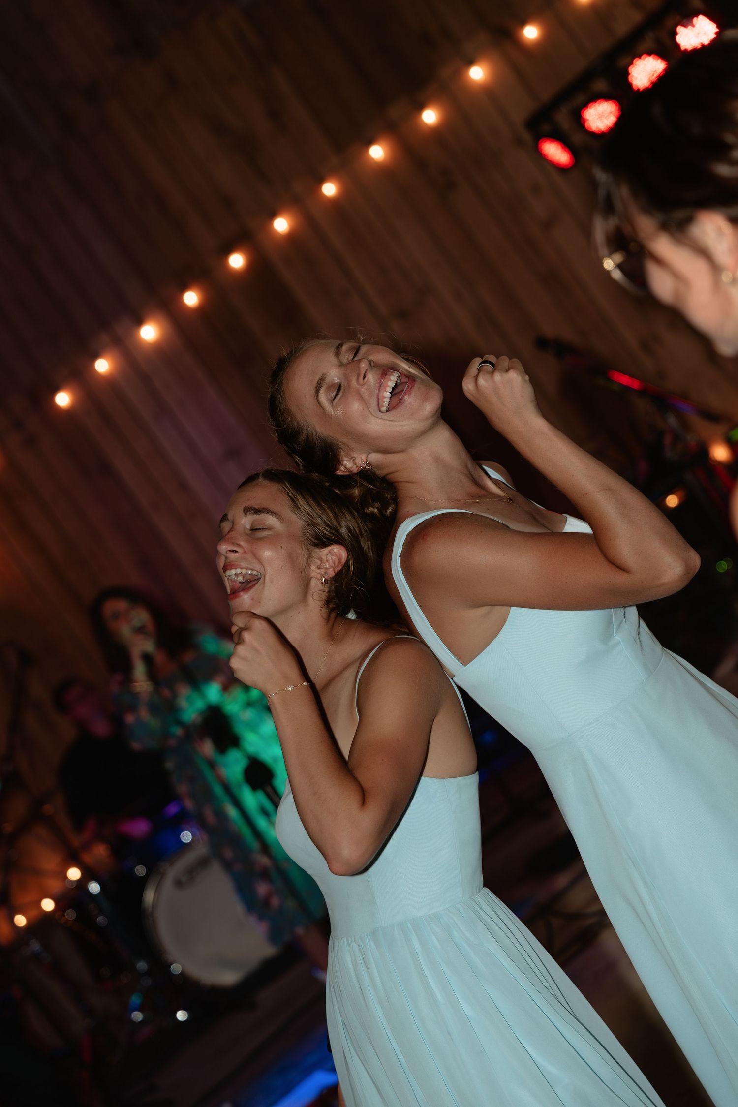 Two bridesmaids in light blue dresses laughing and dancing joyfully at wedding reception with string lights.