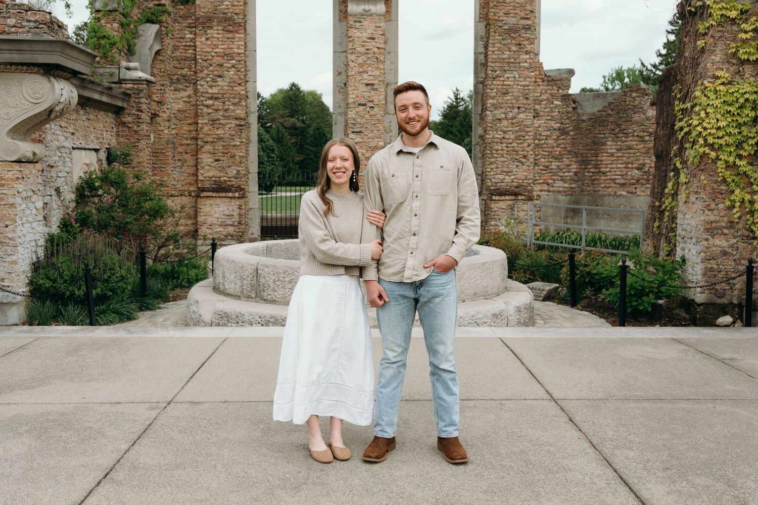 Smiling couple posing together in front of historic stone ruins in a lush garden setting.