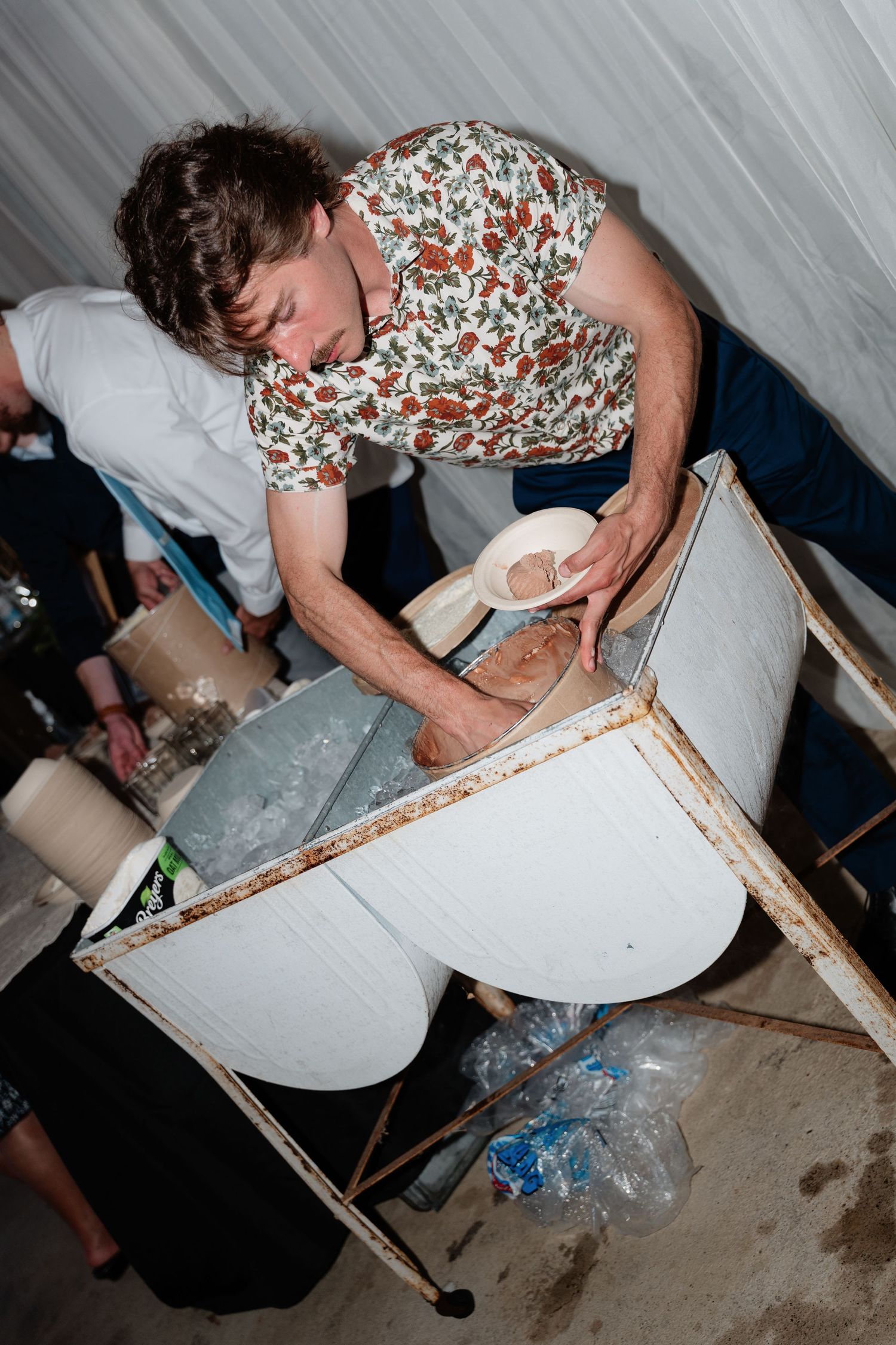 A man in a floral shirt scoops ice from a cooler at a casual outdoor wedding reception.