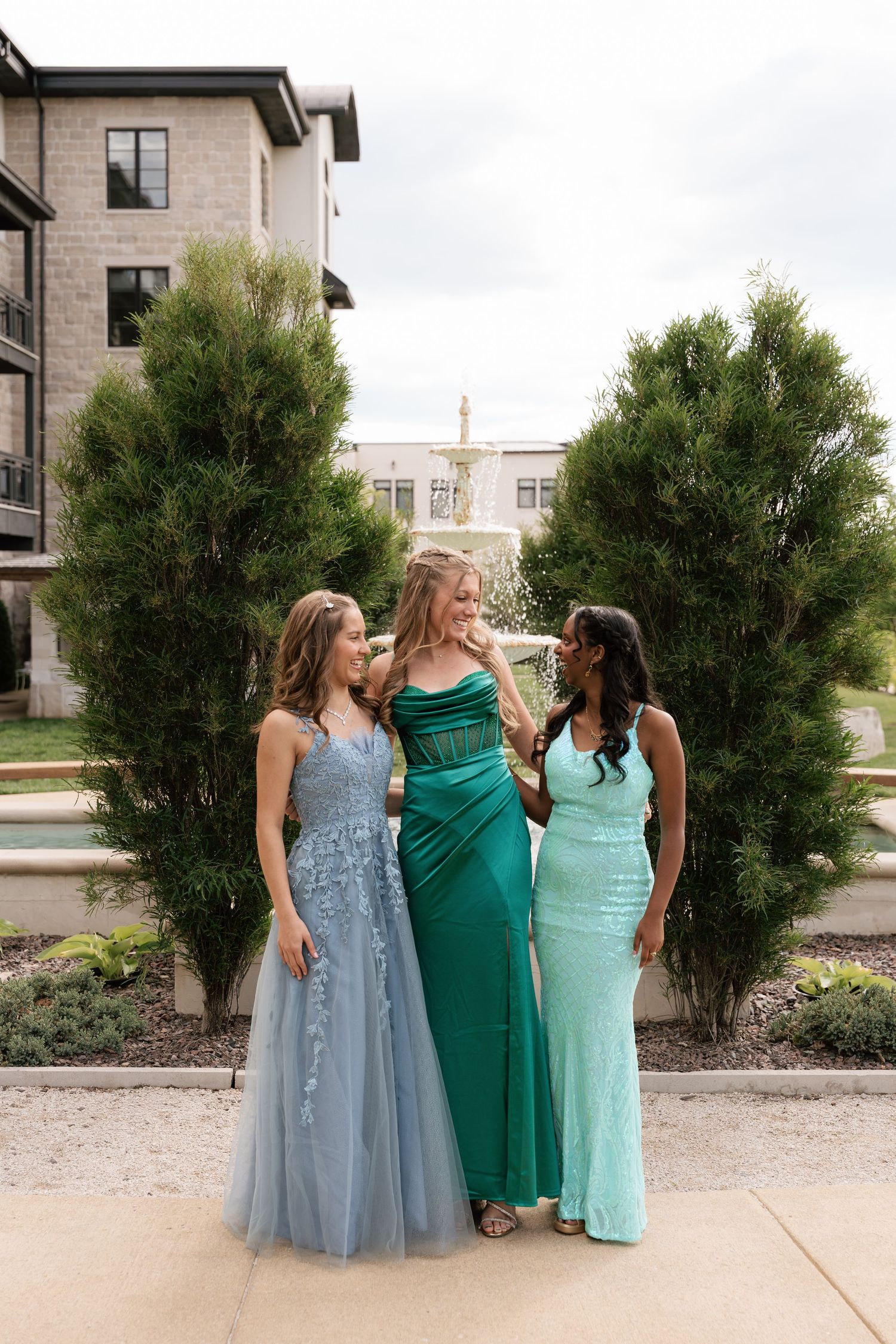 Three young women in elegant formal gowns pose outdoors near a fountain, wearing blue, green, and mint prom dresses.