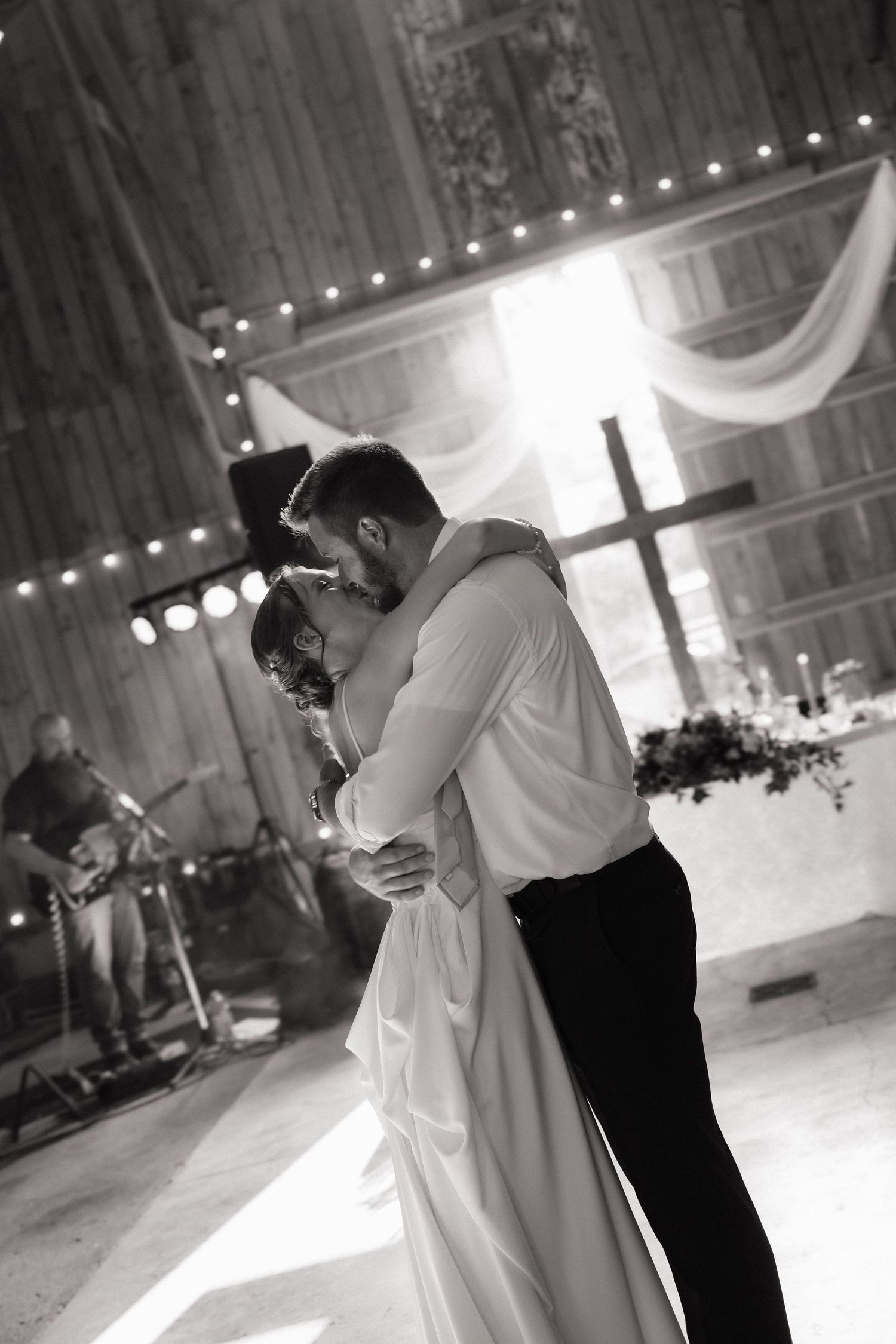 Black and white romantic first dance photo of bride and groom embracing in warmly lit rustic barn venue.