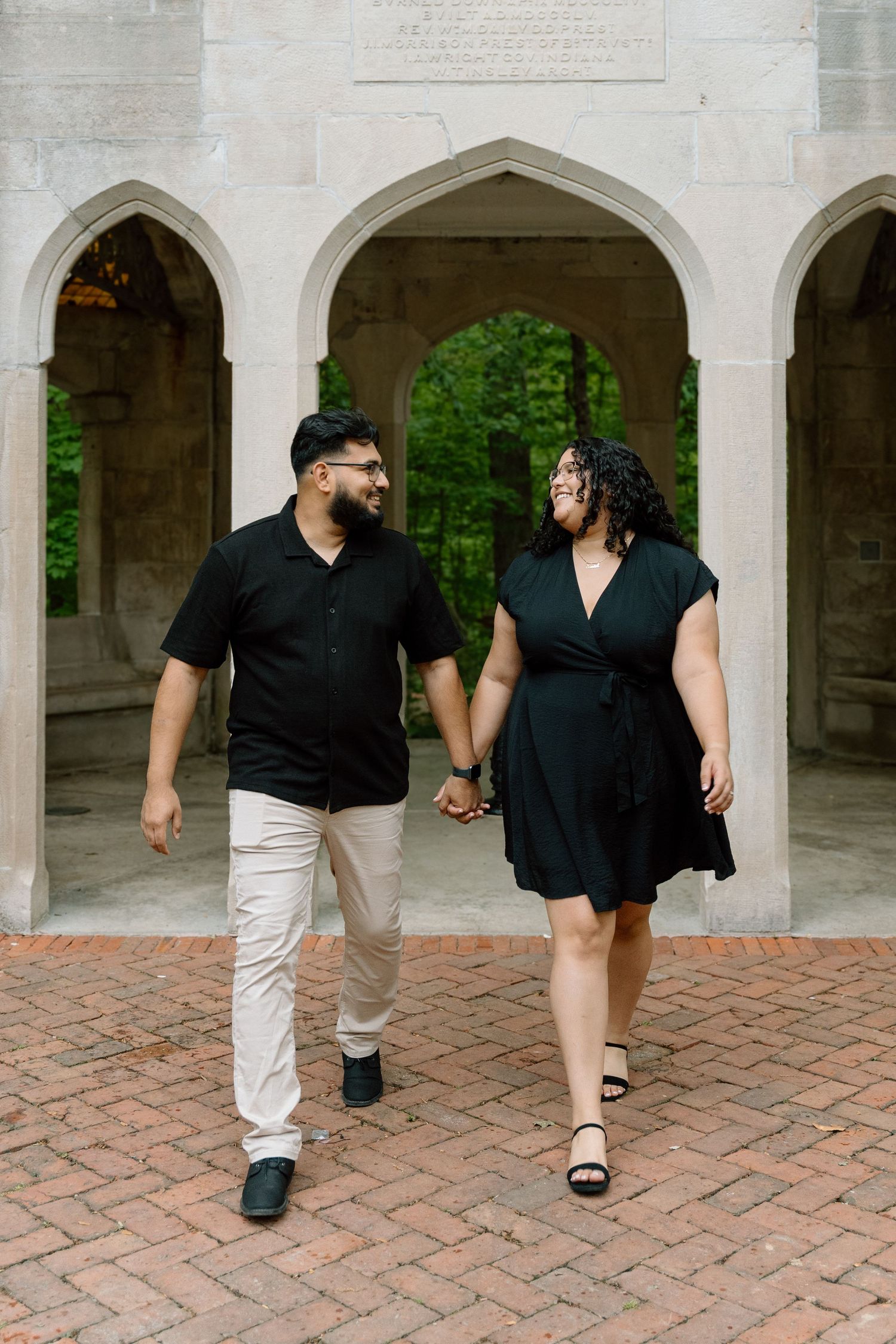 Couple in matching black outfits holding hands and smiling at each other while walking on brick path under stone arches.