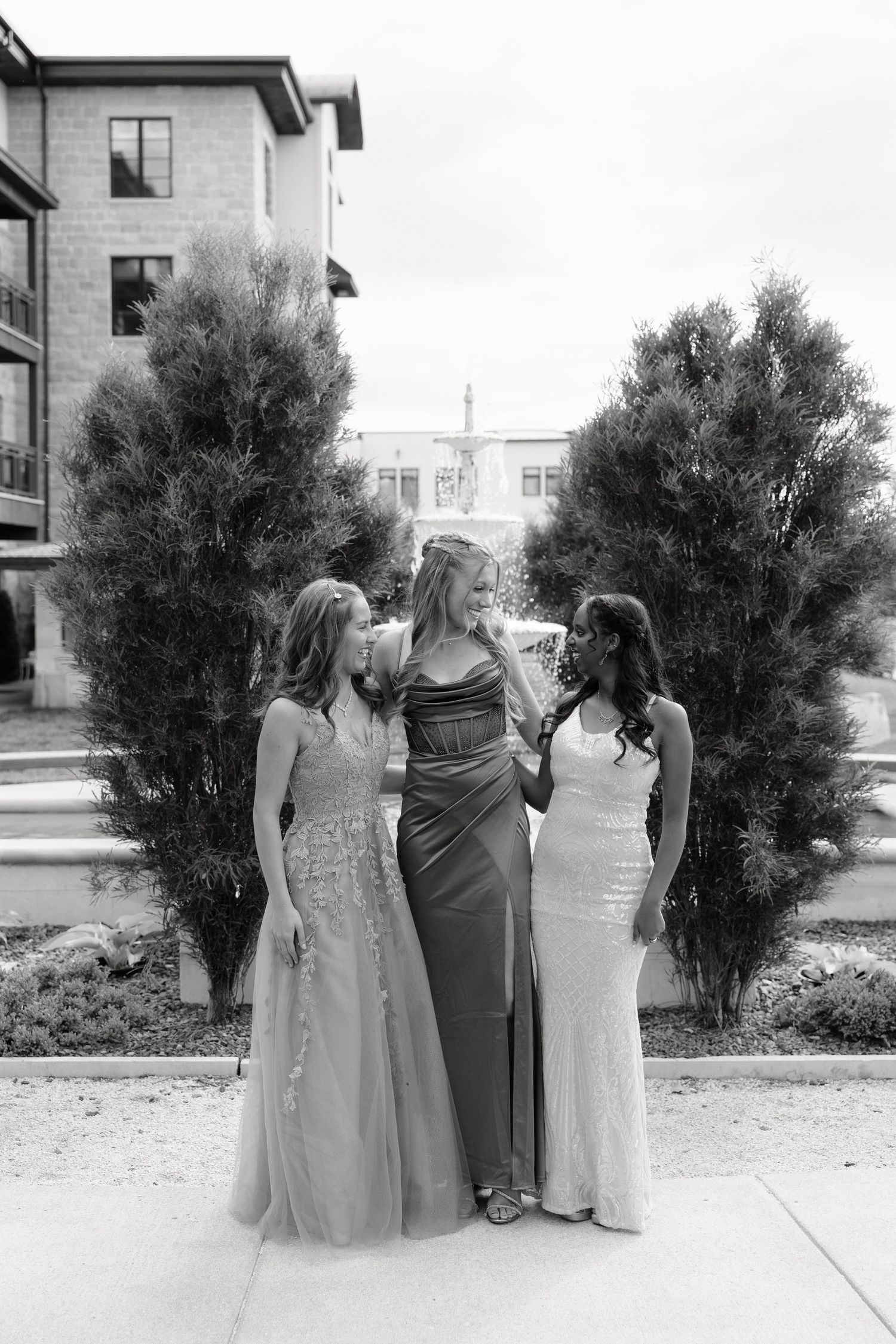 Three young women in elegant formal gowns pose together outdoors near tall shrubs and a fountain in black and white.