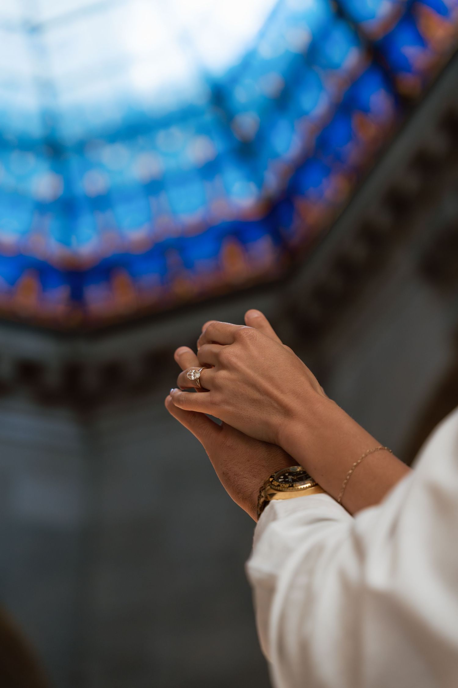 Hands clasped in prayer beneath a stunning blue stained glass dome ceiling in a church.