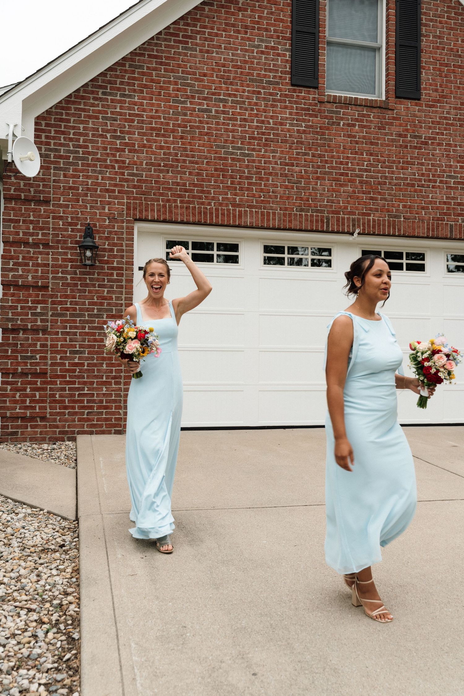 Two bridesmaids in light blue dresses carrying colorful bouquets walk along a driveway beside a brick building.