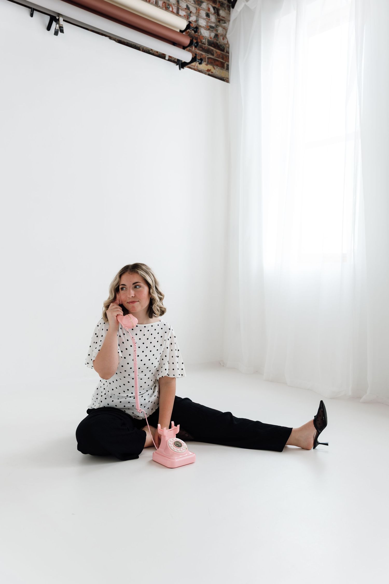 Woman in polka dot blouse sitting on white studio floor talking on a vintage pink rotary telephone.