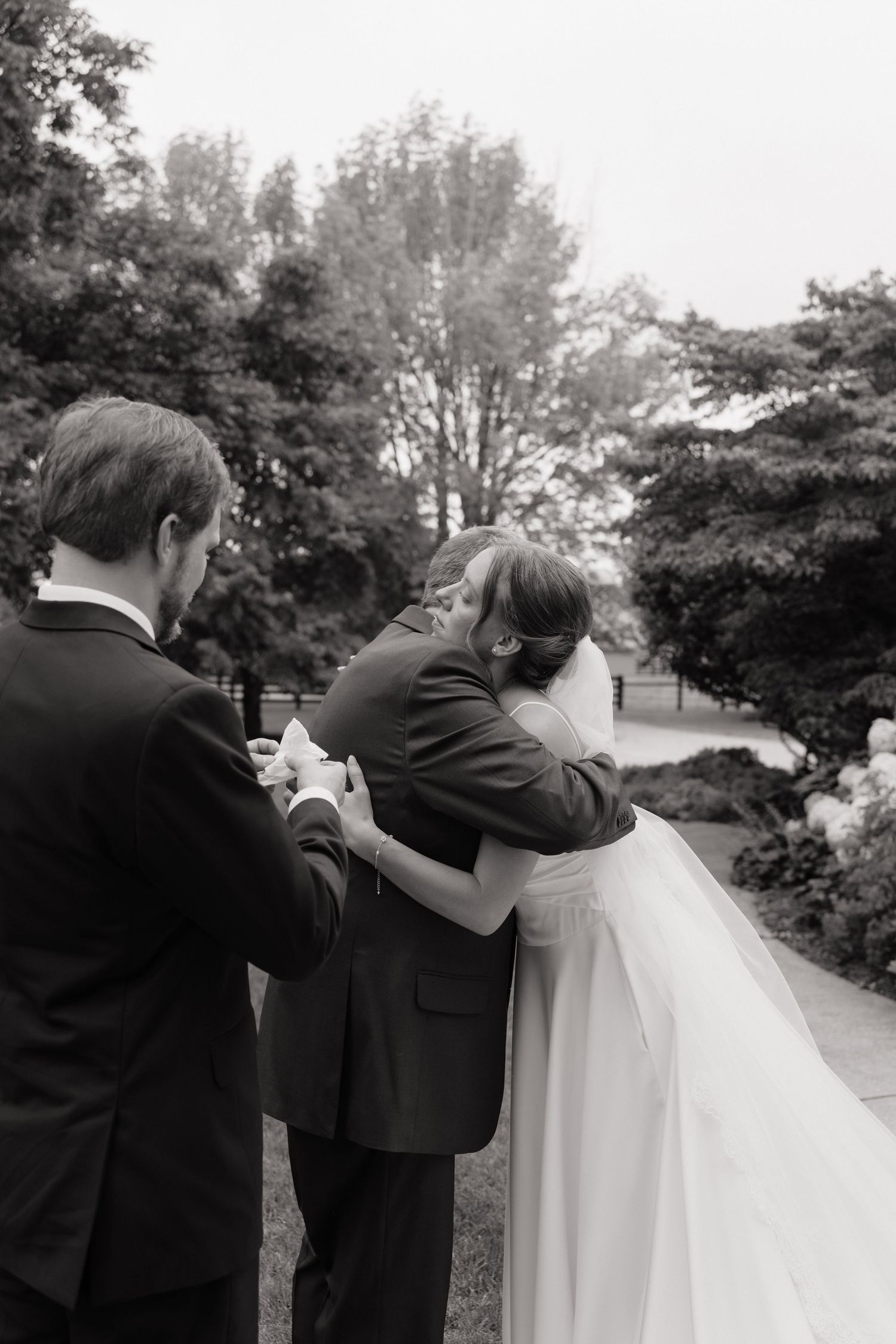Black and white photo of bride embracing family member with groom nearby in lush garden setting.