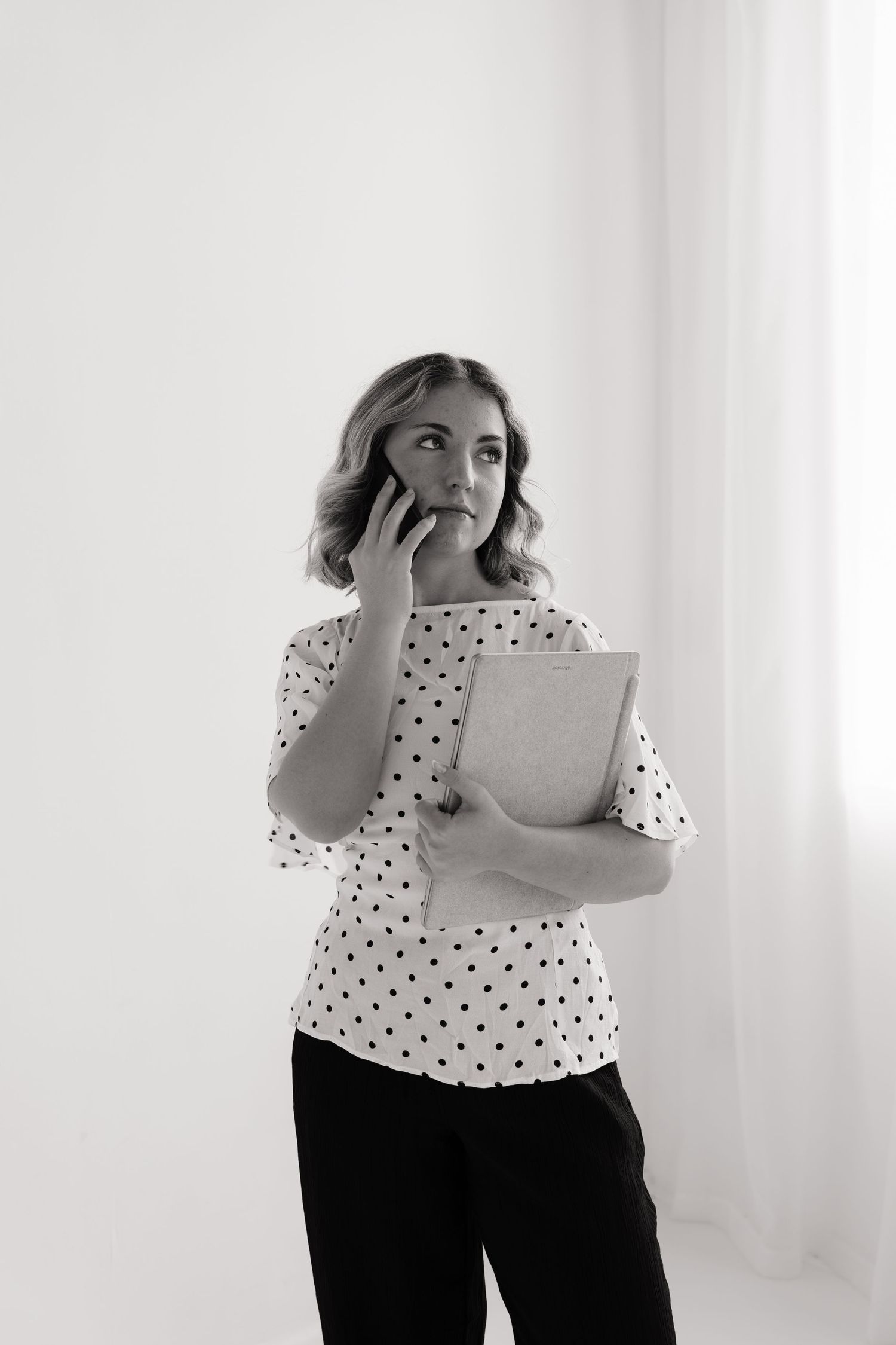 Professional woman in polka dot blouse holding folder while talking on smartphone in black and white photo.
