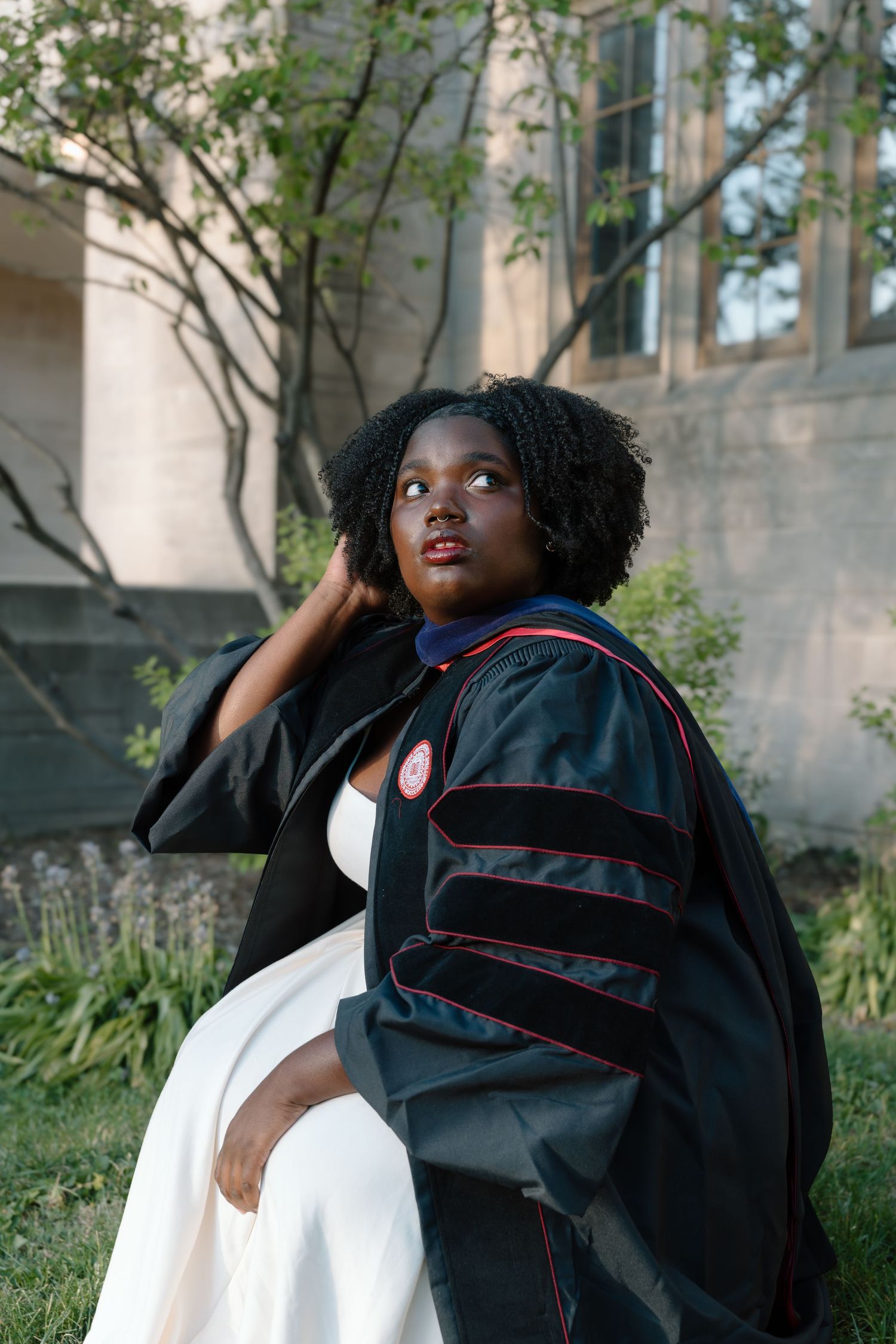 Young woman in doctoral graduation gown and white dress sits outdoors on grass near a stone building.