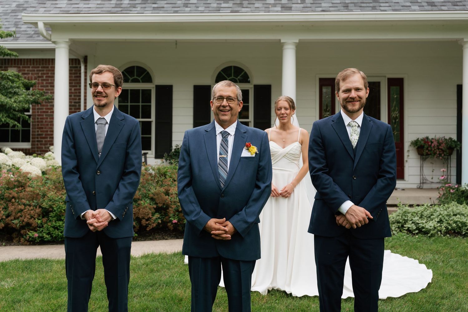 Wedding party with bride in white gown and three men in navy suits posing outdoors in front of a brick house.
