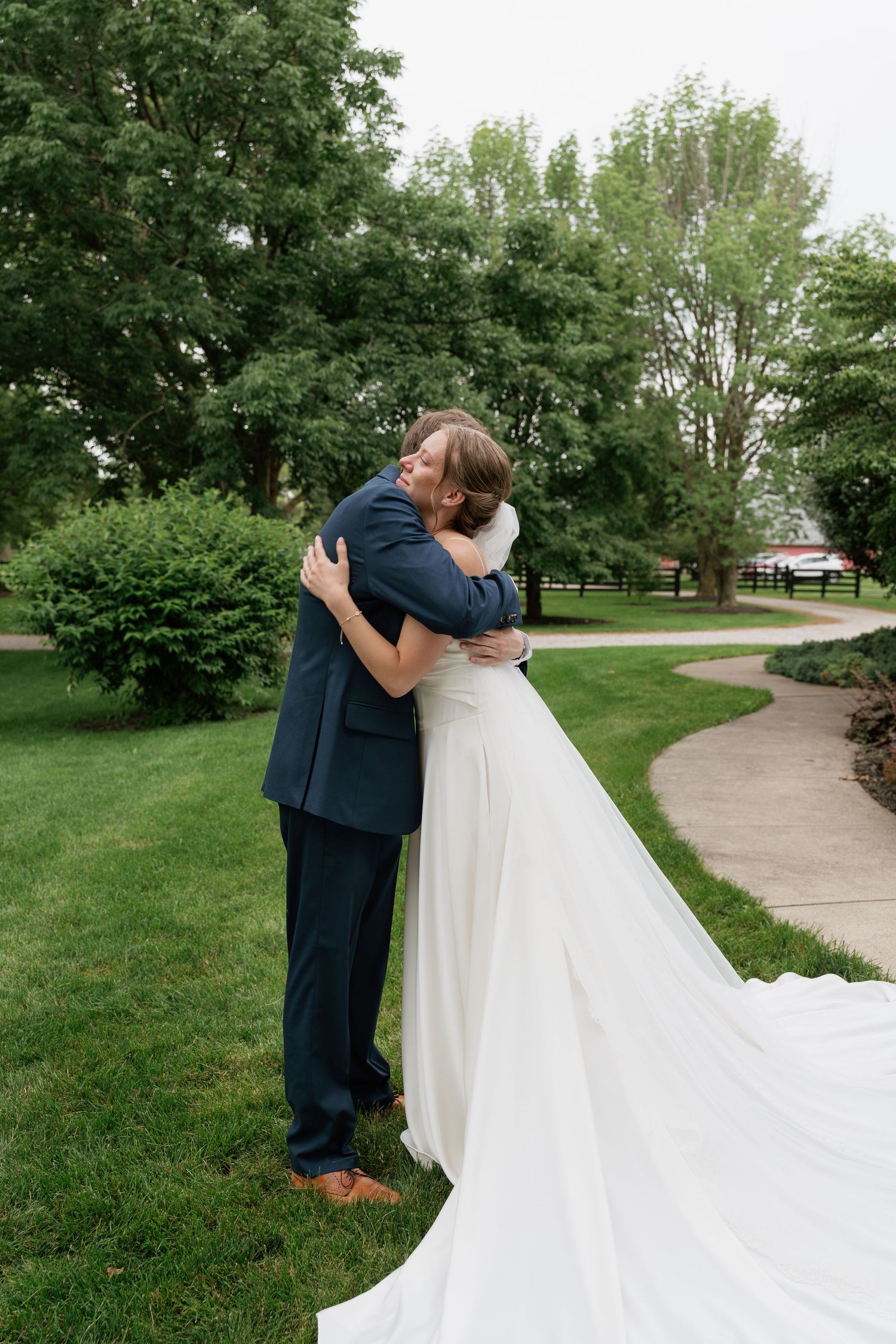 Bride and groom share a heartfelt hug outdoors on their wedding day, surrounded by lush green trees and gardens.