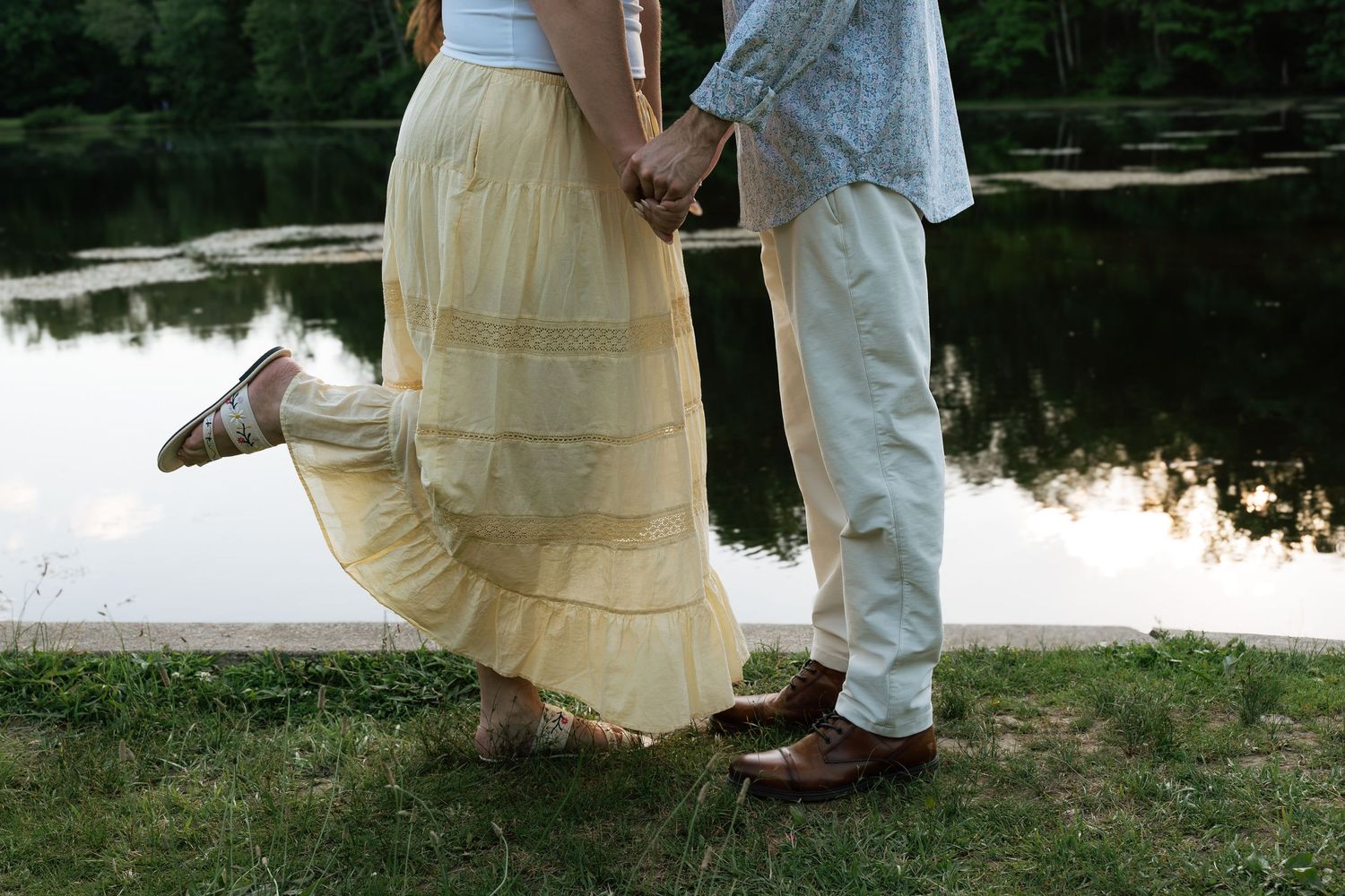 Couple holding hands by a serene lake, woman in yellow maxi skirt and man in light pants on green grass.