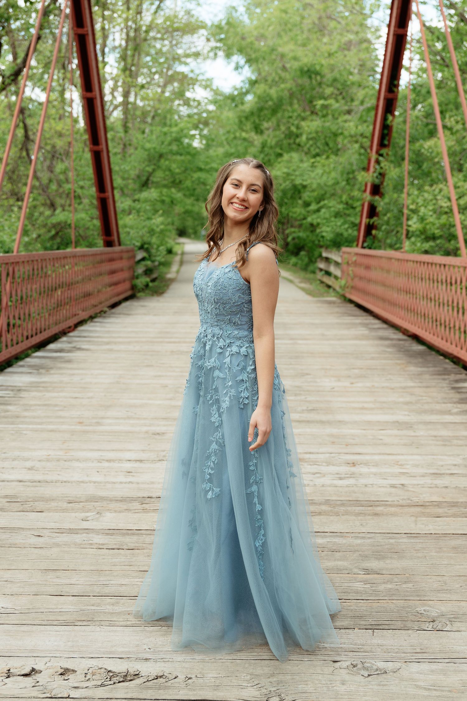Young woman smiling in a blue floral tulle prom gown on a wooden bridge surrounded by lush green trees.