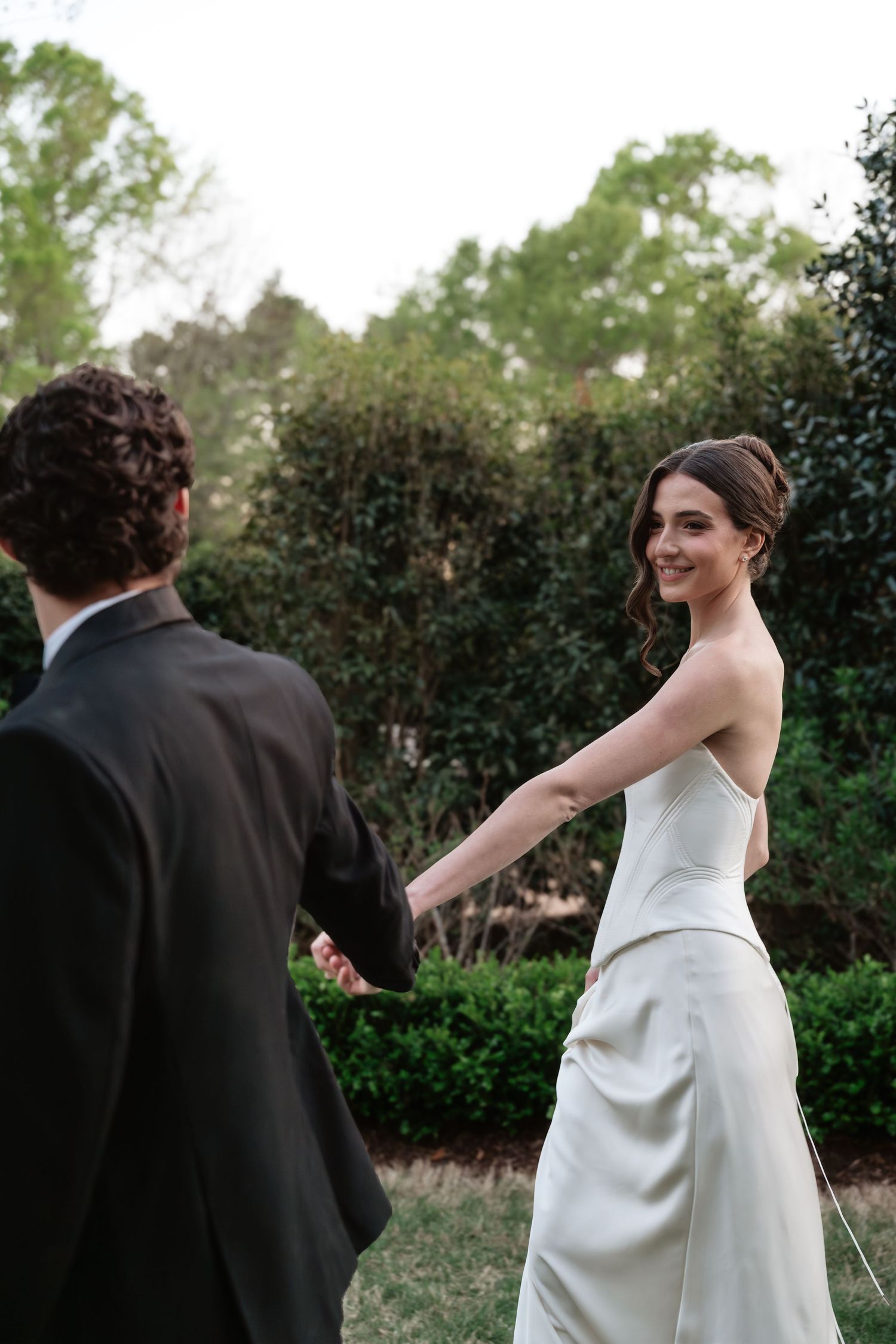 Smiling bride in strapless white gown holds groom's hand outdoors, glancing back with lush green hedges behind them.