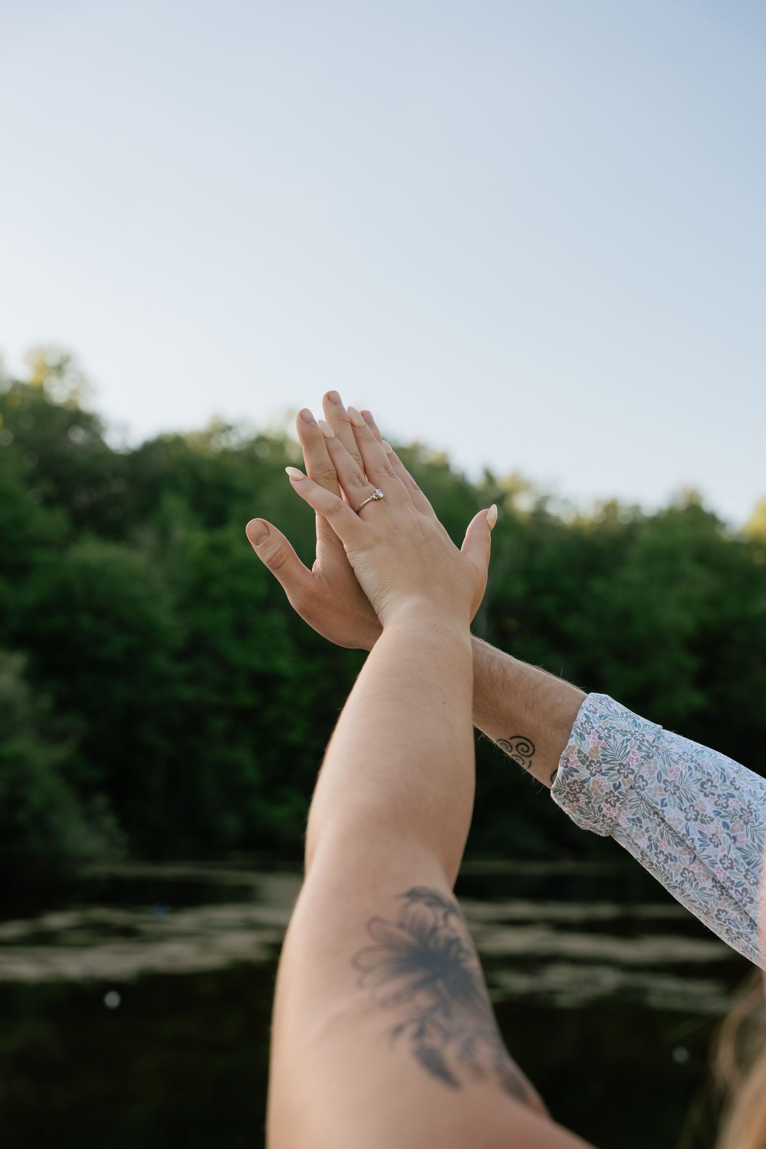 Two hands touching against a green forest backdrop, one wearing a ring and the other with a palm tree tattoo.