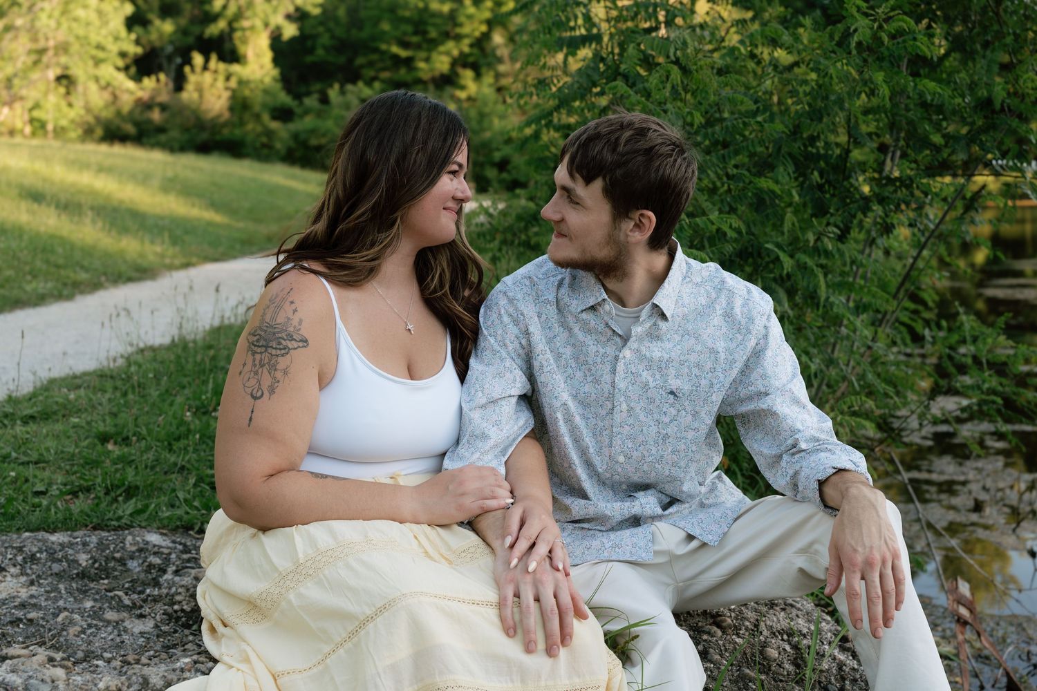 Couple sitting on stone steps outdoors, smiling at each other, woman with tattoo in white crop top and yellow skirt.