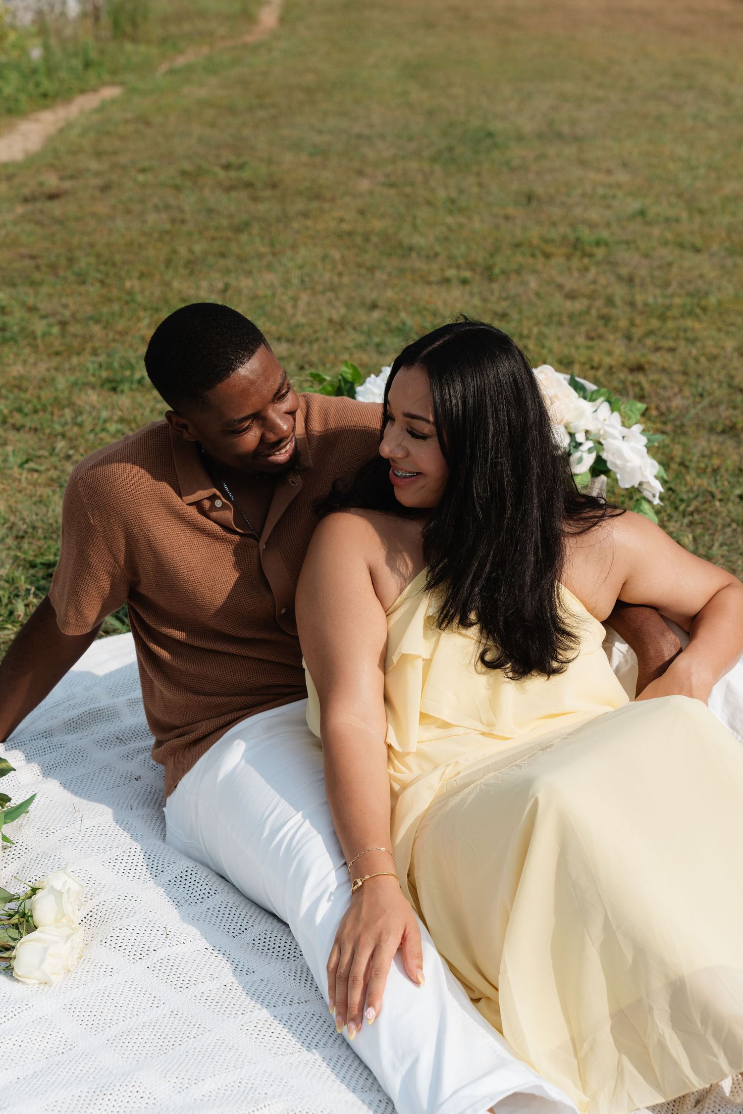 A couple relaxing on a white blanket outdoors, woman in yellow dress leaning against smiling man in brown shirt.