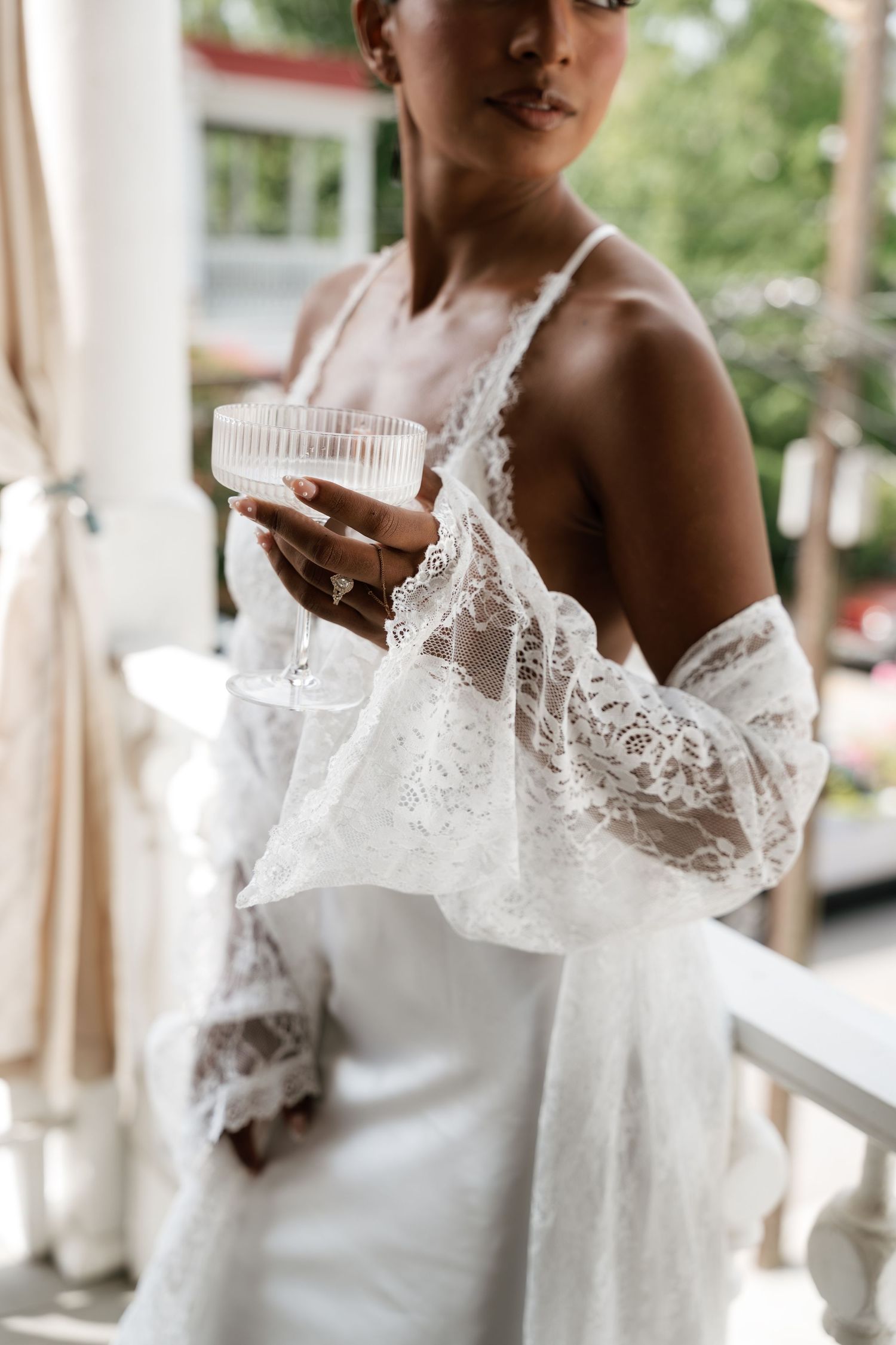Bride in white lace robe and slip dress holding a coupe glass on a sunlit porch.
