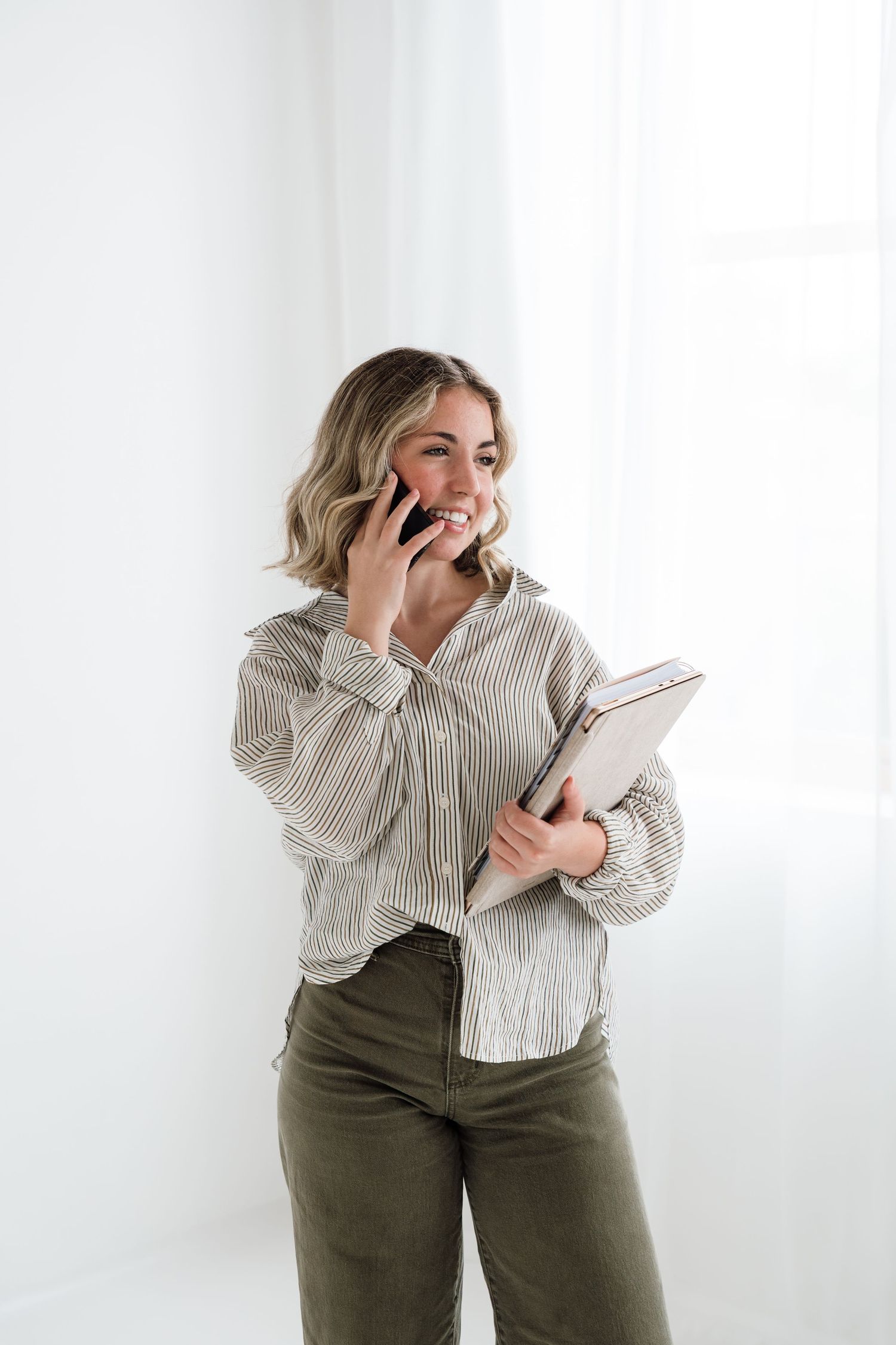 Smiling blonde woman in striped shirt talking on phone while holding a notebook against white background.