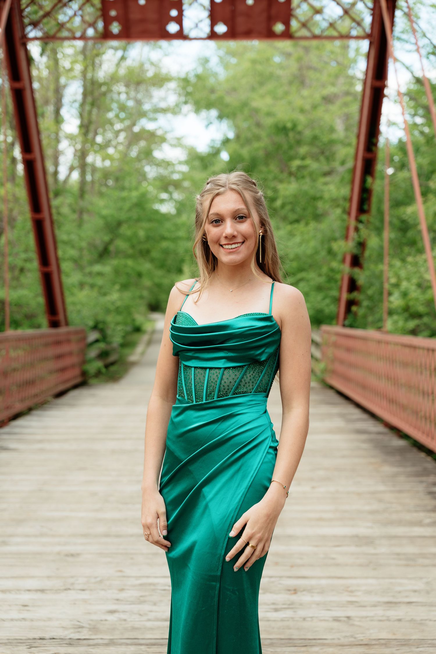 Young woman in elegant emerald green satin gown posing on a rustic red iron bridge surrounded by lush greenery.