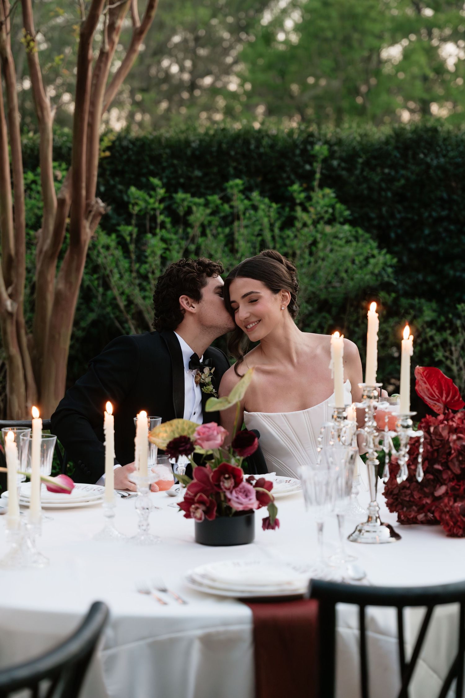 A bride and groom share a romantic moment at a candlelit outdoor wedding reception table adorned with red floral arrangements.