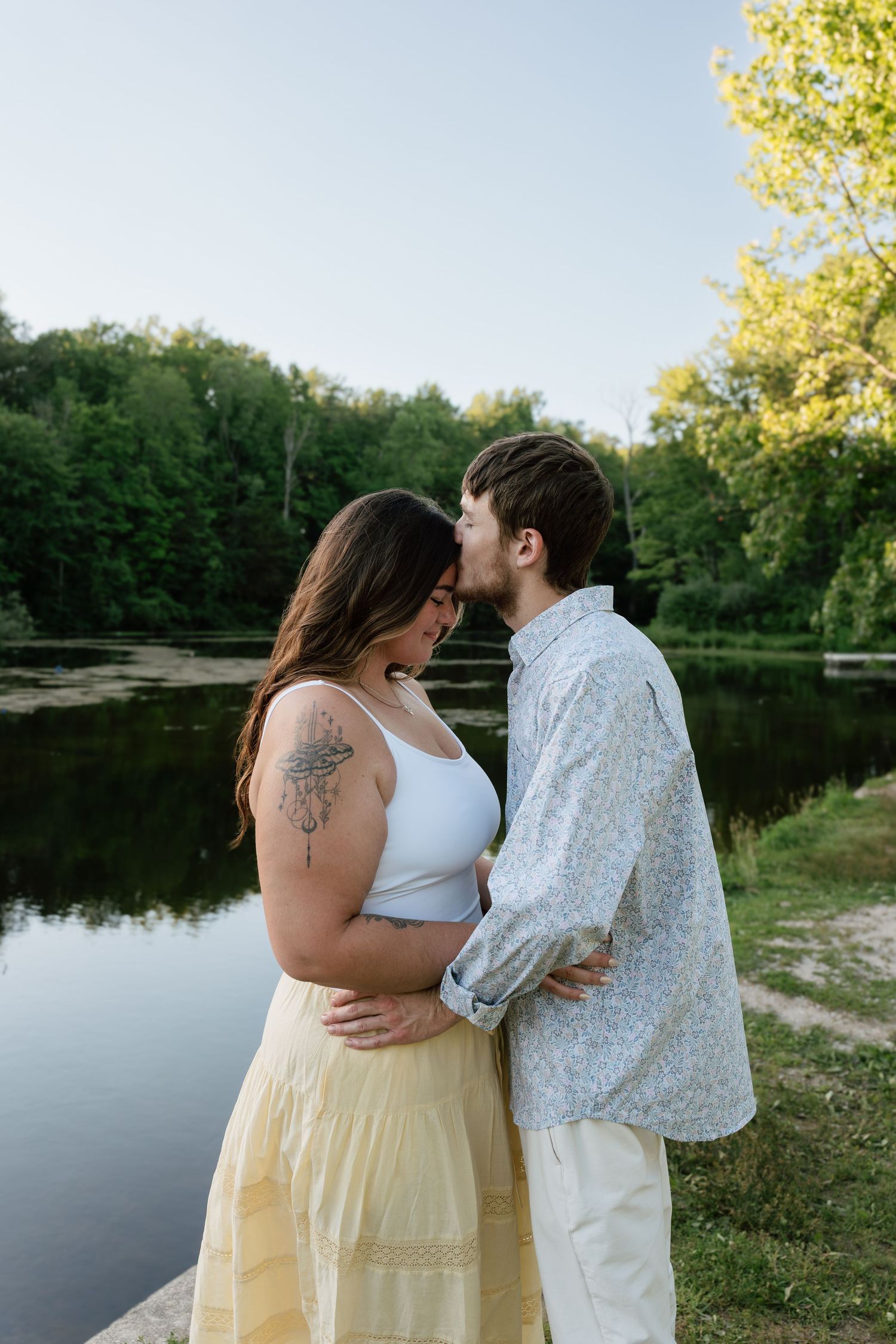 A couple shares a tender kiss by a serene lake surrounded by lush green trees during golden hour.