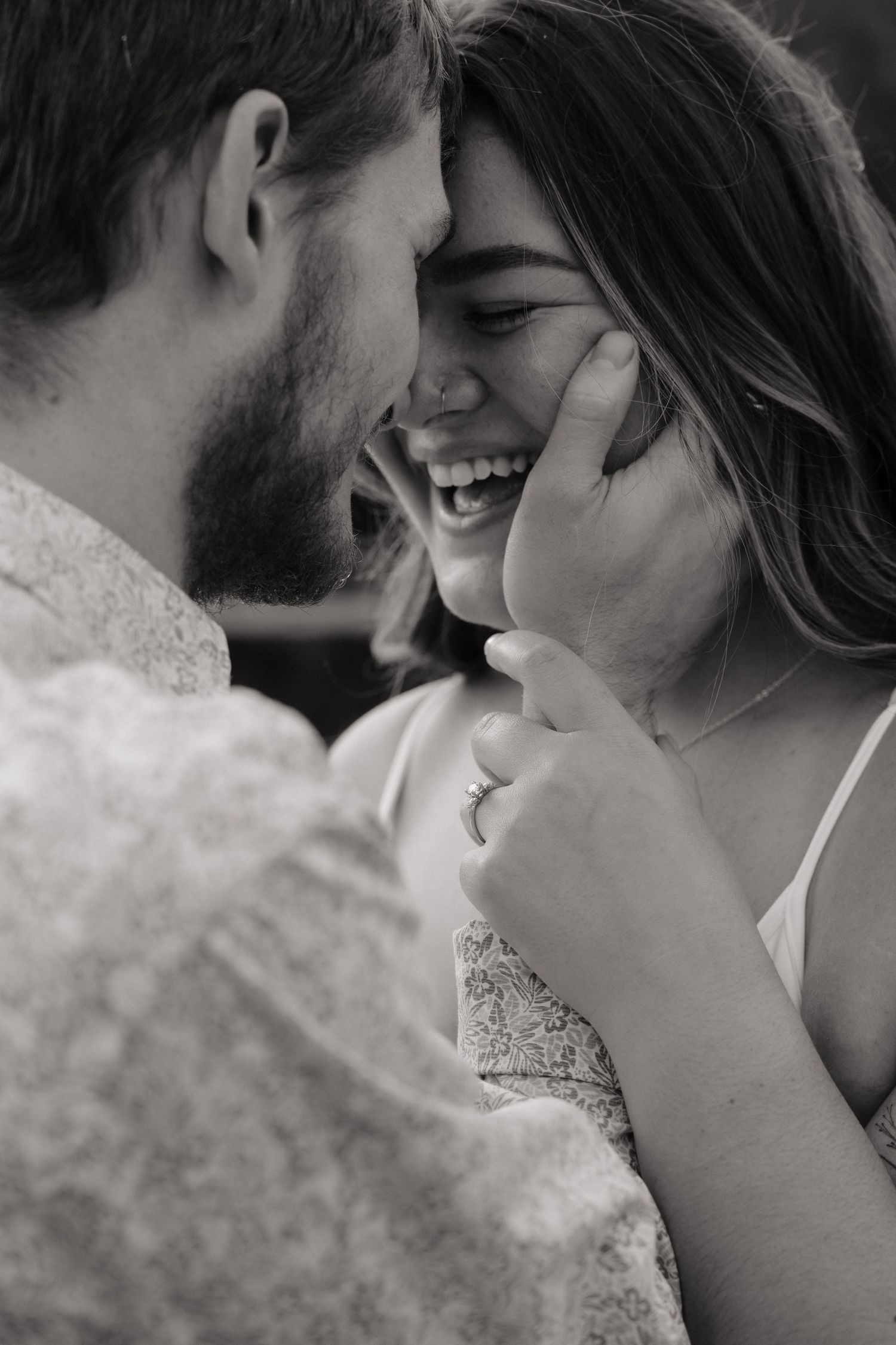 Black and white photo of a laughing couple sharing an intimate moment, touching foreheads, woman wearing an engagement ring.