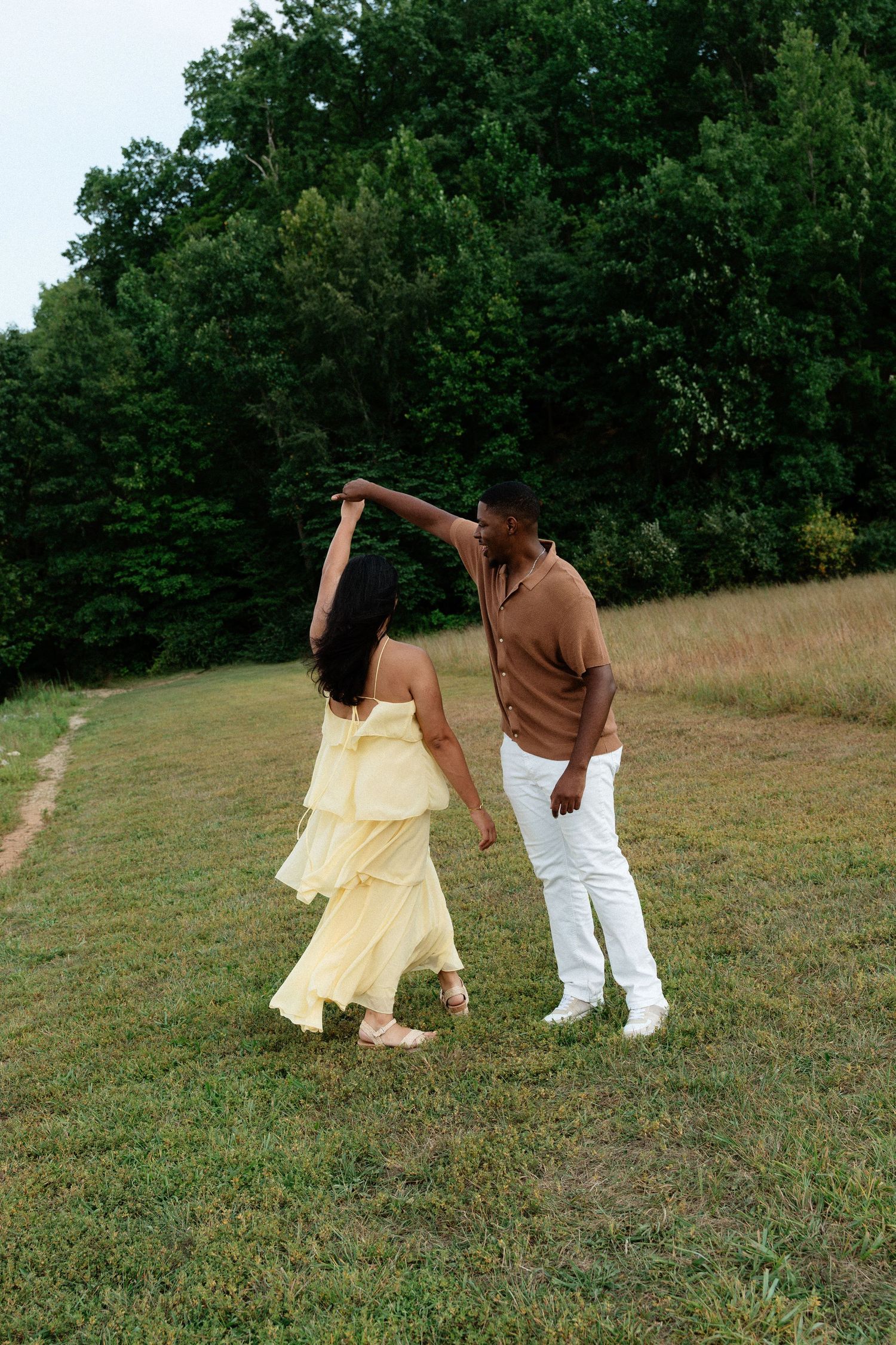 A couple dancing in a green field, woman in yellow dress being twirled by man in white pants and brown shirt.