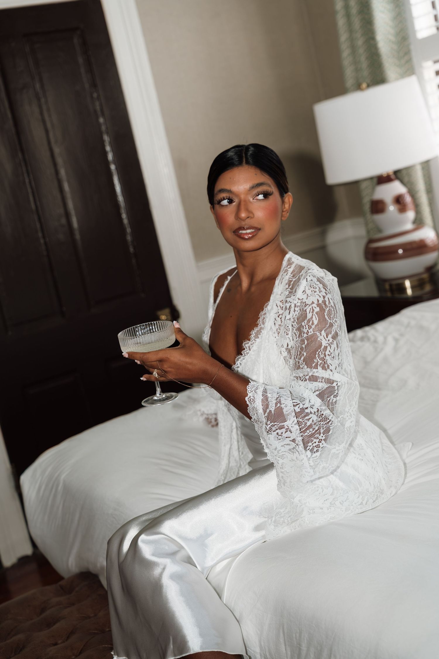 Elegant bride in white lace robe holding a champagne coupe glass while sitting on a bed in a luxury hotel room.