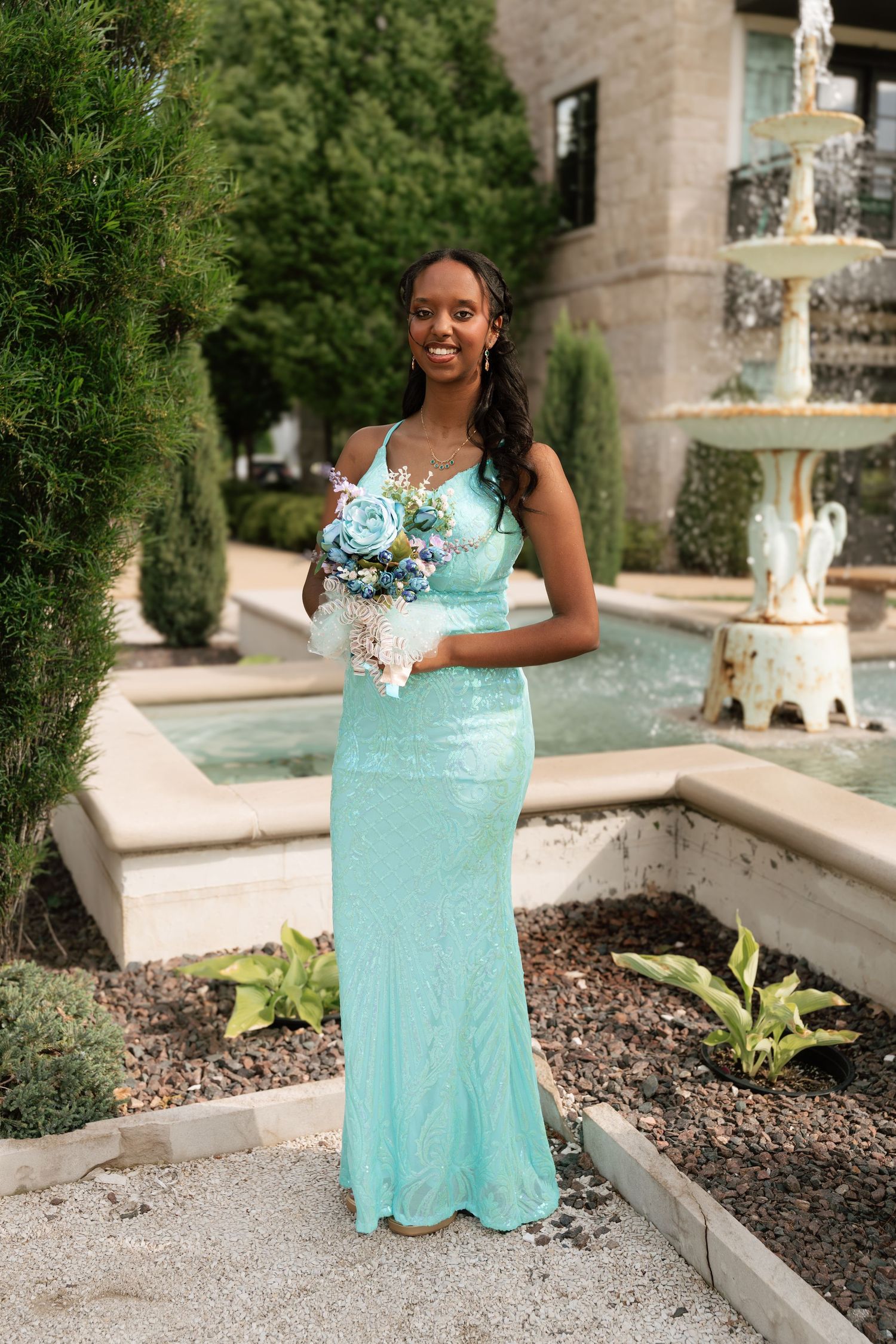 Young woman in a mint green sequin gown holds a blue bouquet near a stone fountain outdoors.