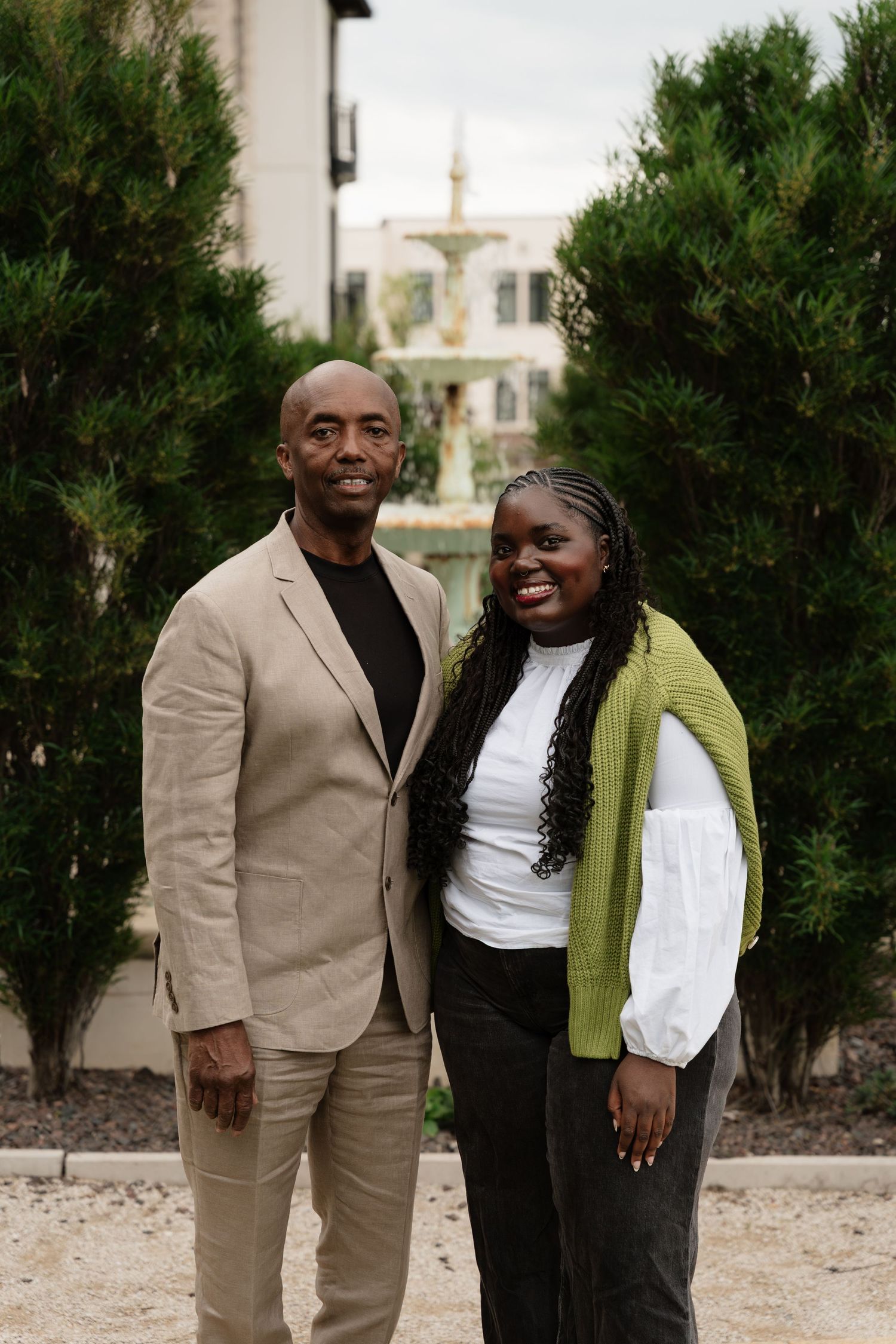 A man in a beige blazer and a woman in a green vest and white blouse smile together outdoors near lush green trees.