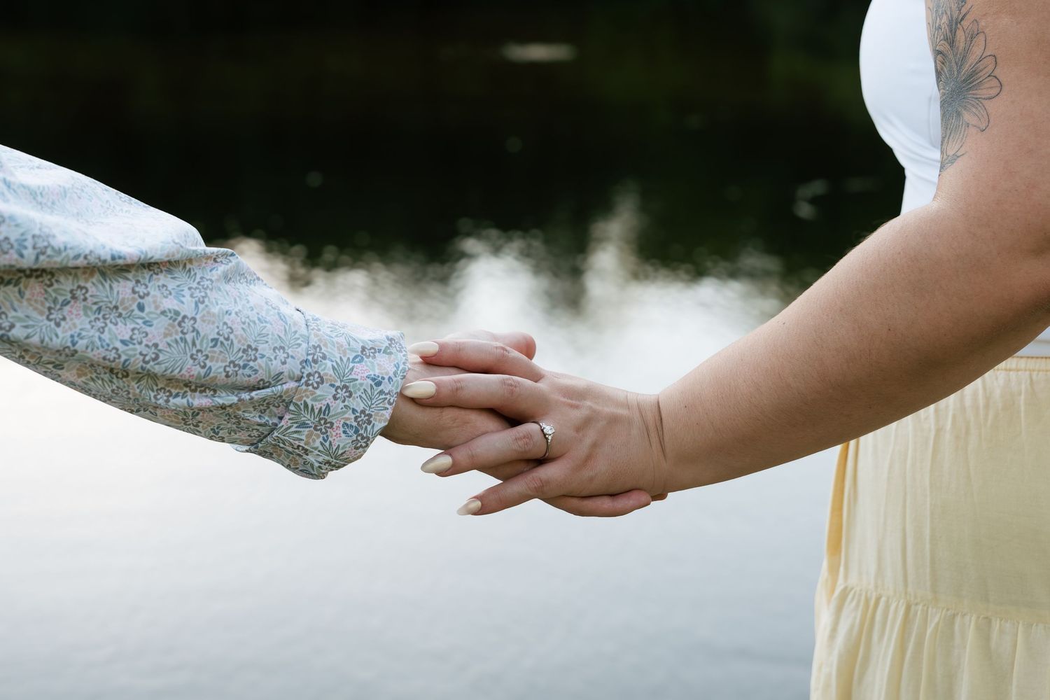 Two women holding hands near a serene lake, one wearing a floral sleeve and the other a yellow dress with a tattoo.