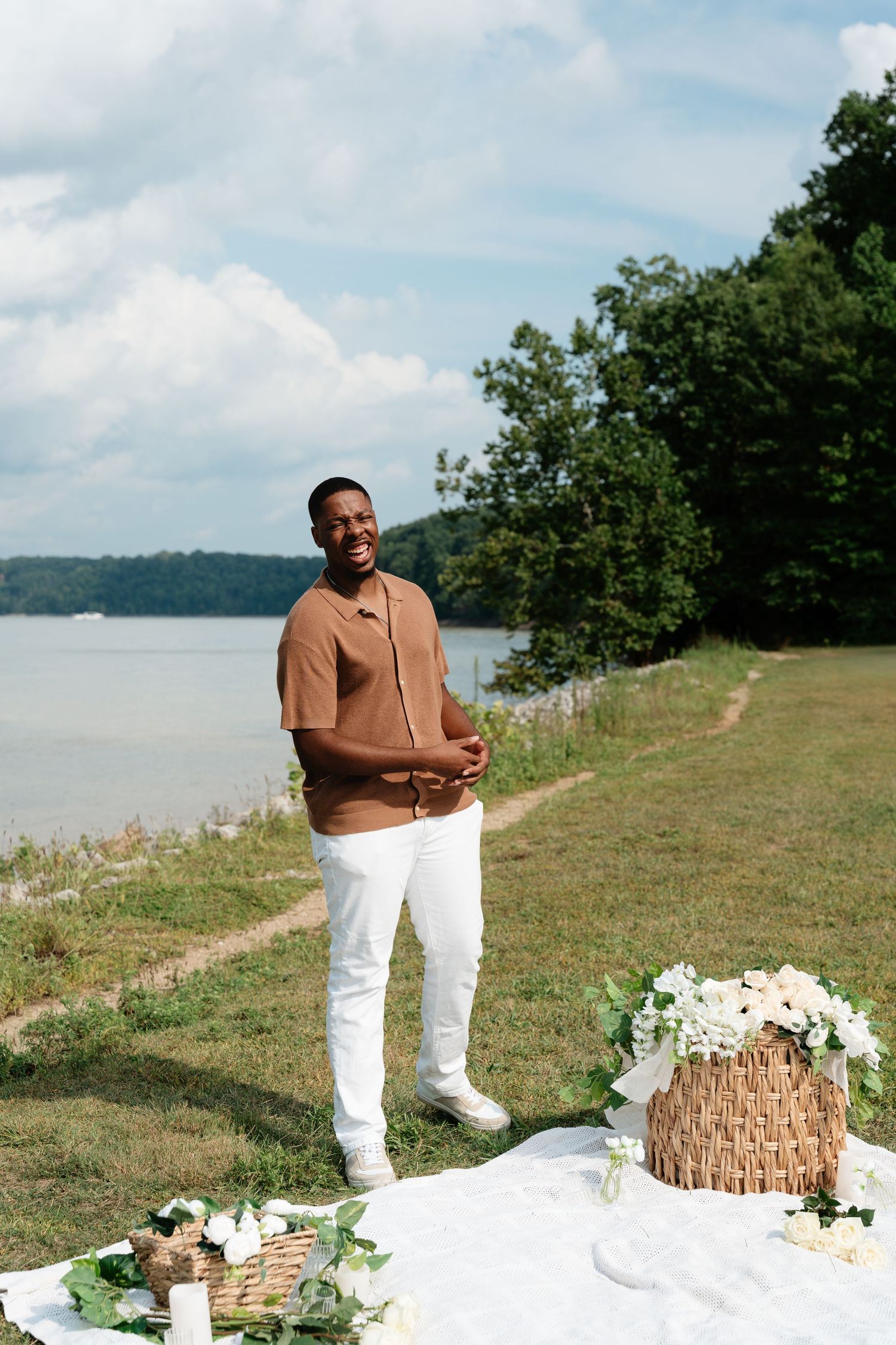 Smiling man in brown shirt and white pants stands near a lakeside picnic setup with flowers and a wicker basket.