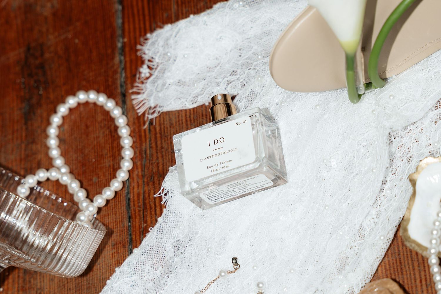 Elegant flat lay of I Do perfume bottle with pearl bracelet, white lace fabric, and nude heels on wood.