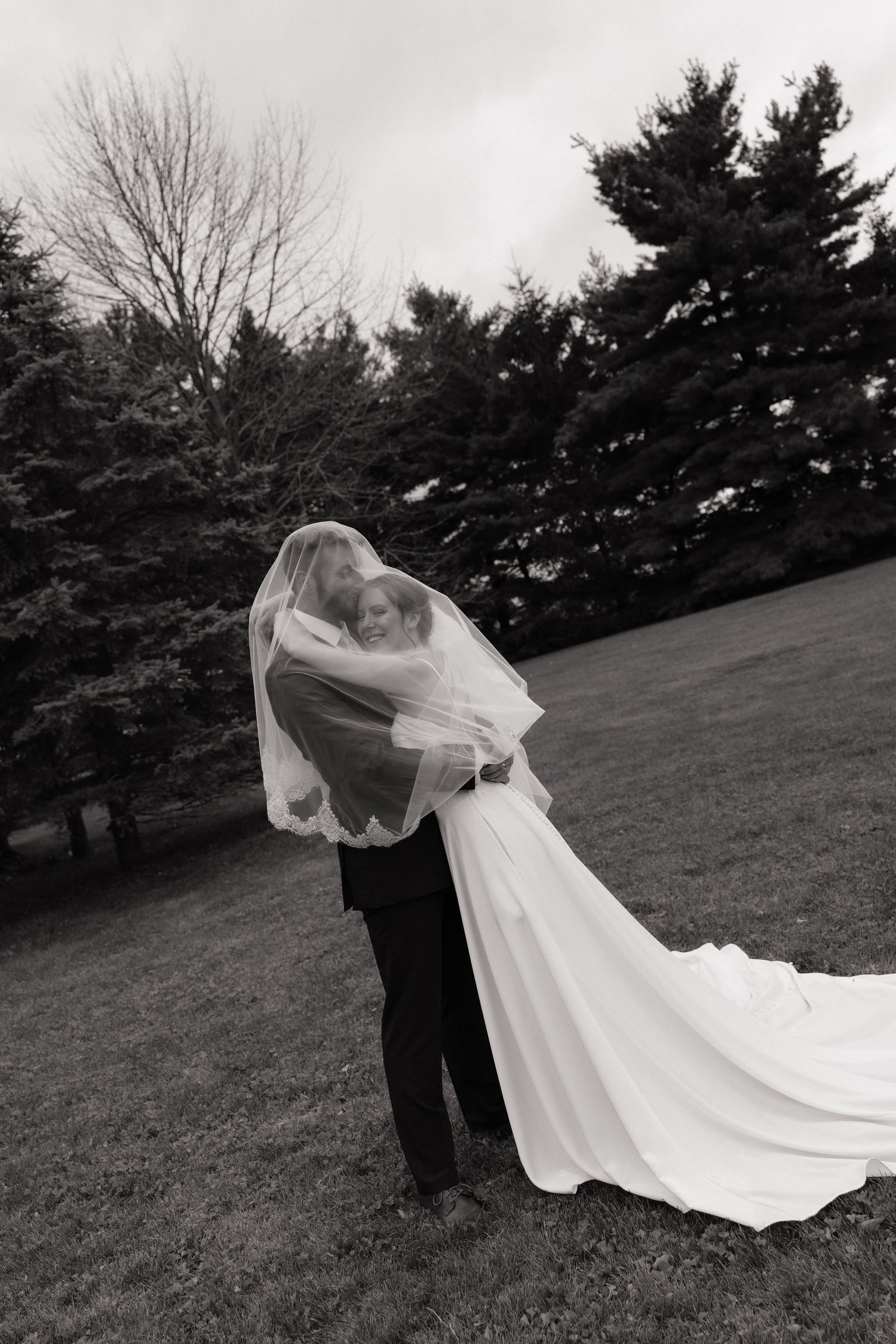Black and white photo of a bride and groom kissing under a flowing veil outdoors on their wedding day.