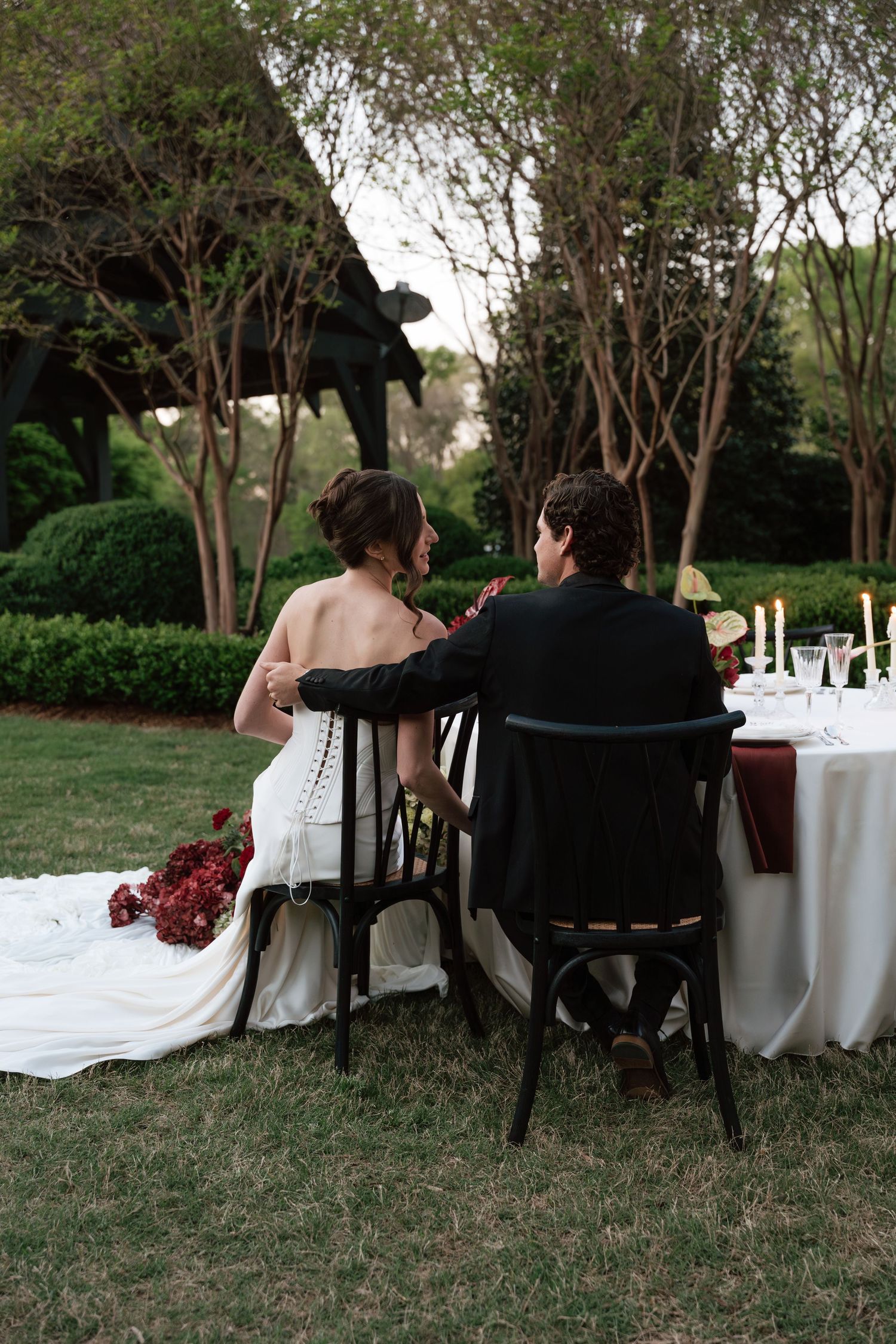 Bride and groom seated at elegant outdoor wedding reception table surrounded by lush greenery and candlelight.