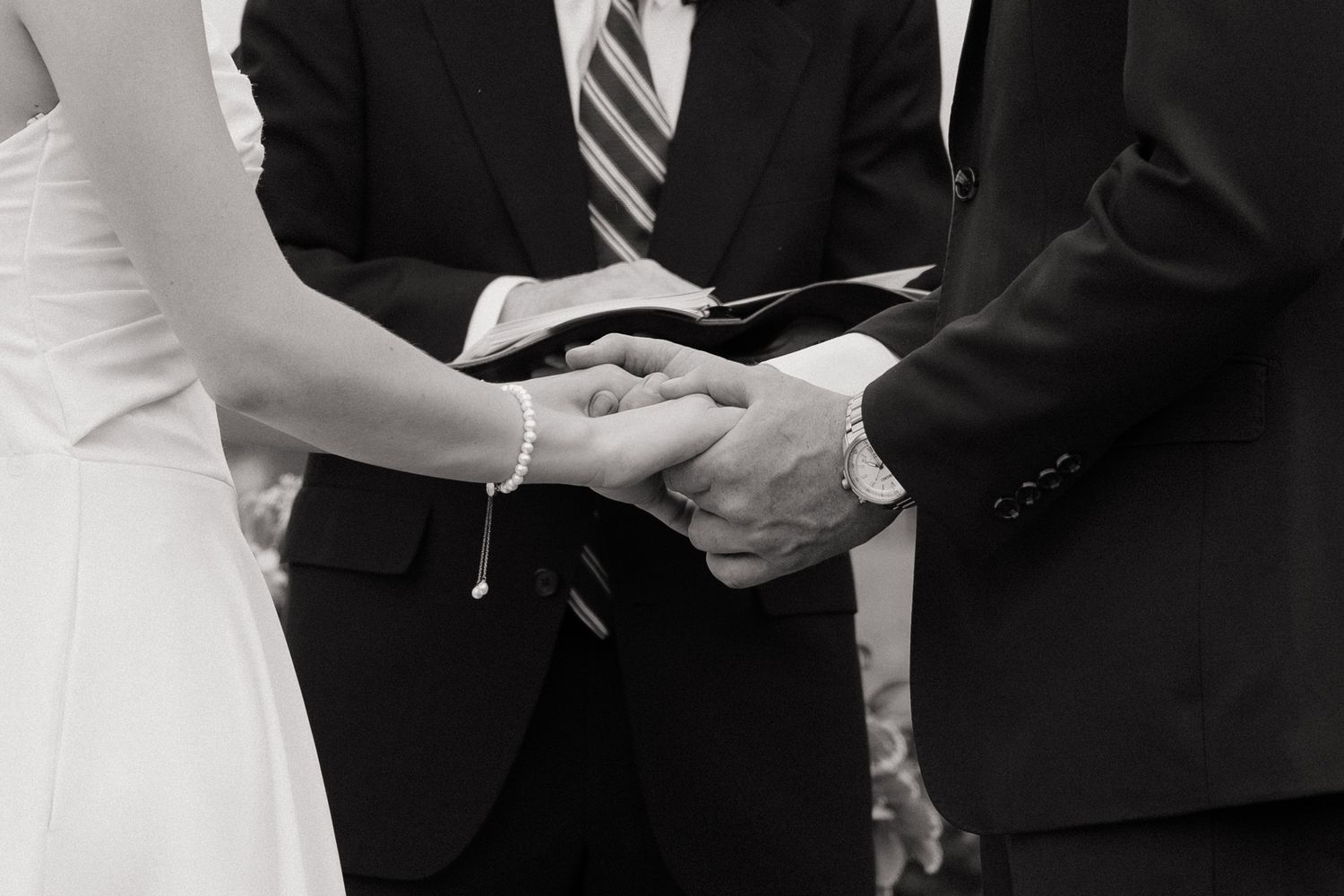 Black and white wedding ceremony photo of bride and groom holding hands with officiant holding a book.