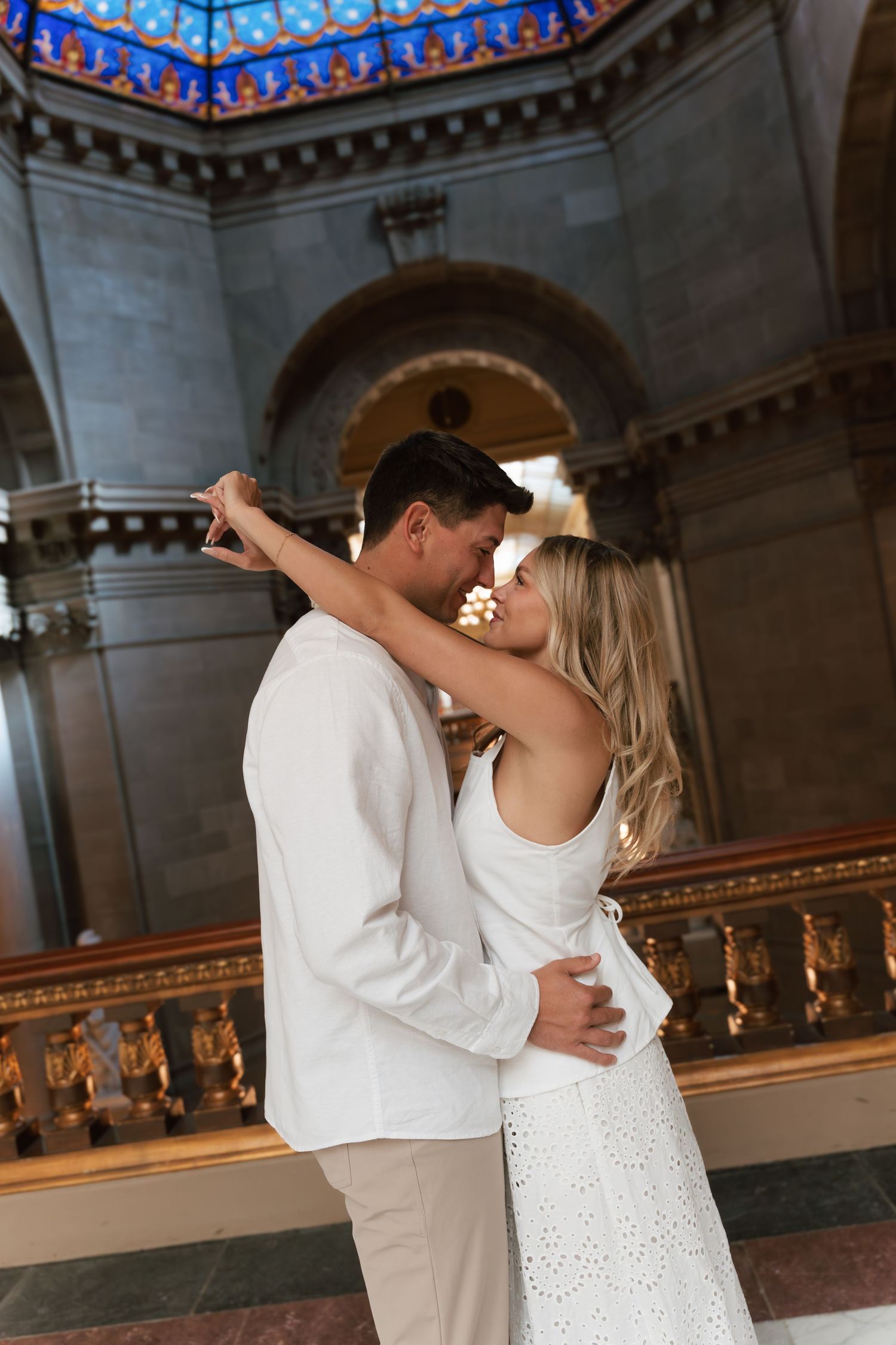 Romantic couple in white outfits embracing and almost kissing inside an elegant domed building with ornate architecture.