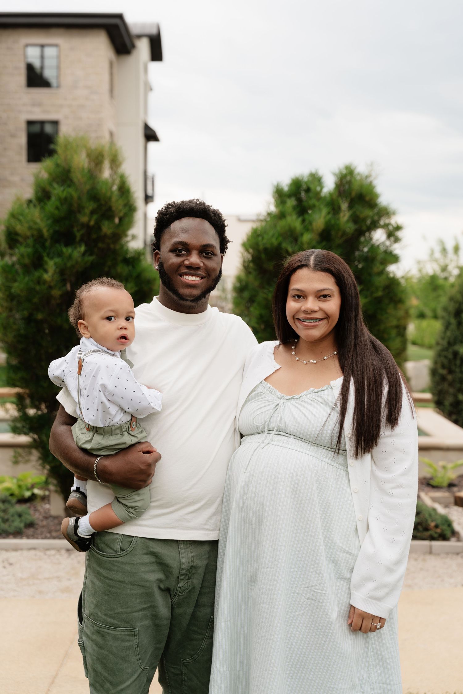 A smiling family poses outdoors, with a man holding a baby beside a pregnant woman in a light blue dress.