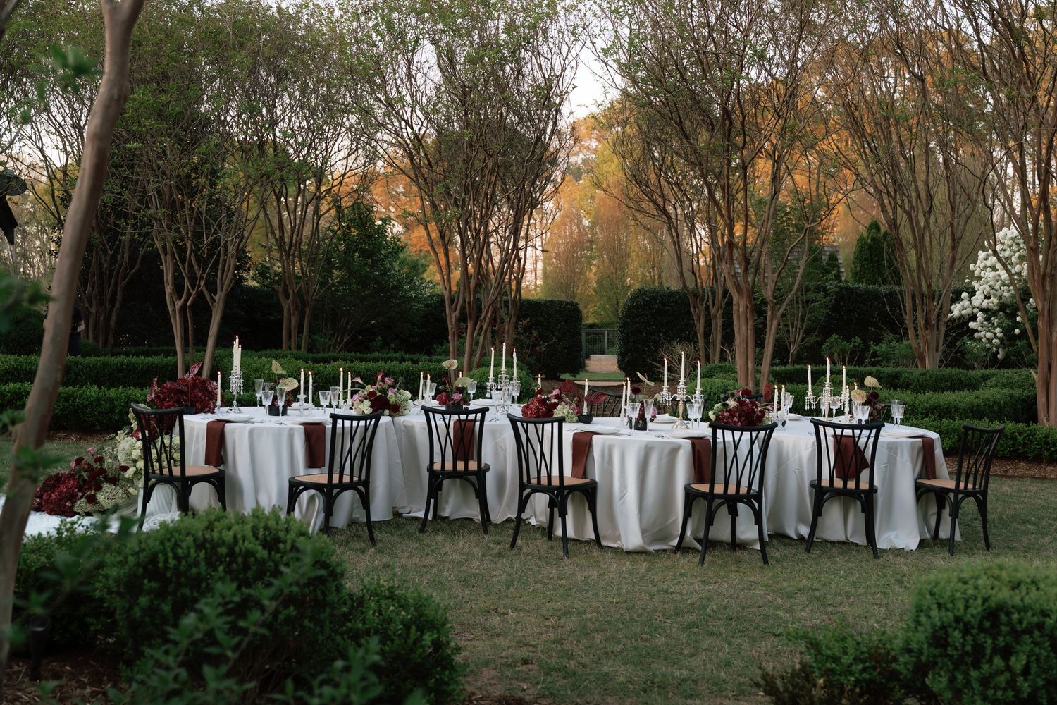 Elegant outdoor wedding reception with white tablecloths, dark chairs, and floral centerpieces set among lush green trees at dusk.
